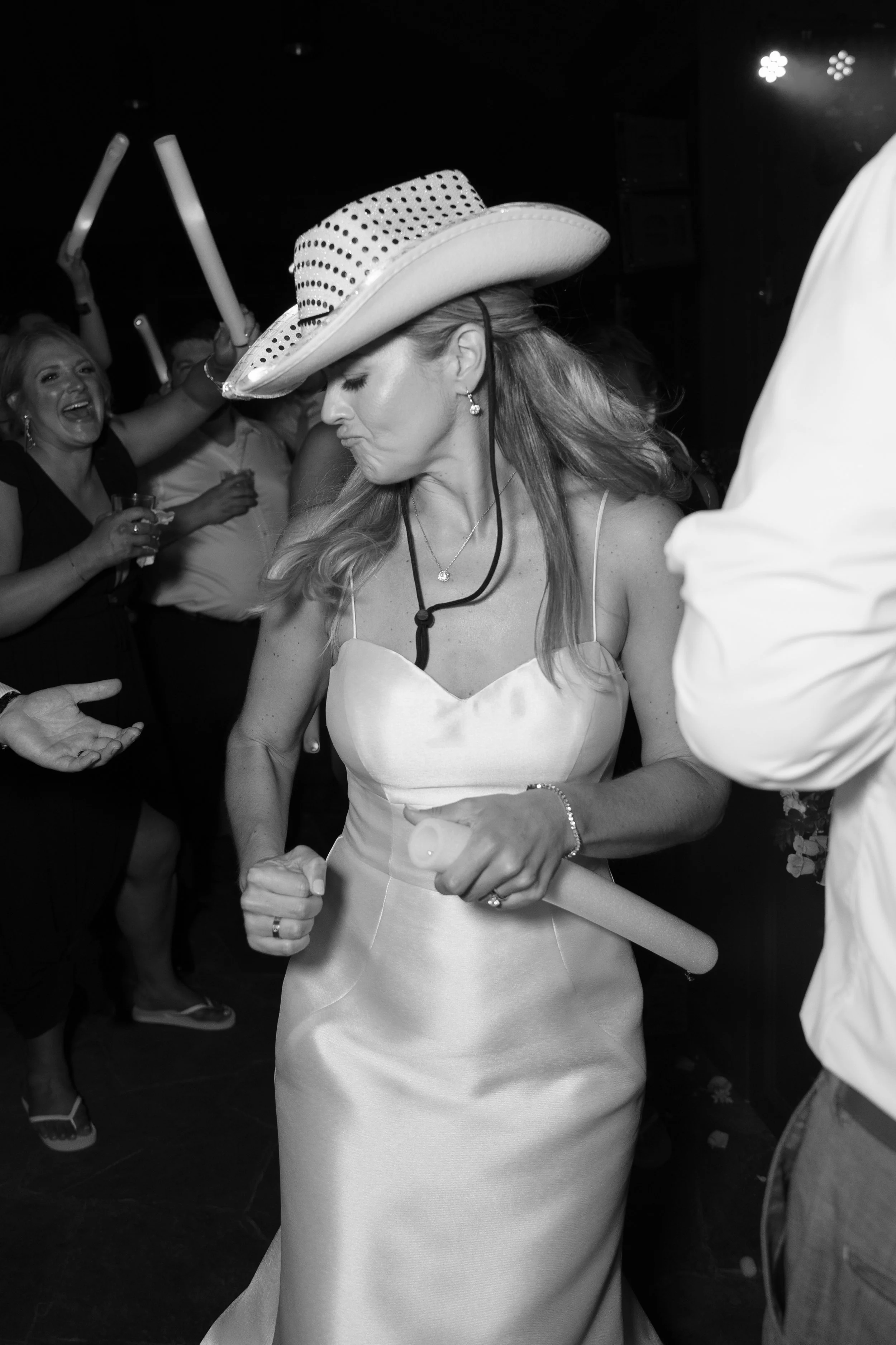 A woman wearing a white satin dress and a large polka dot cowboy hat dances at a party, surrounded by other people.