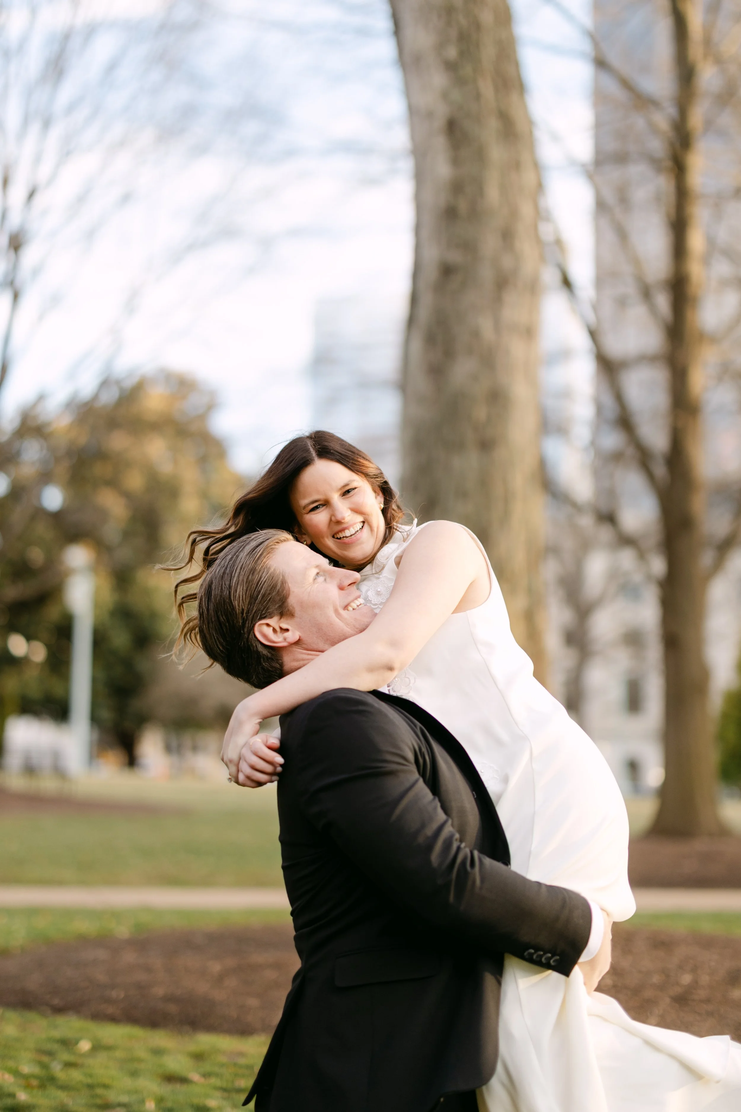 A man in a black suit lifting a woman in a white dress in a park with trees in the background, both smiling and looking happy.