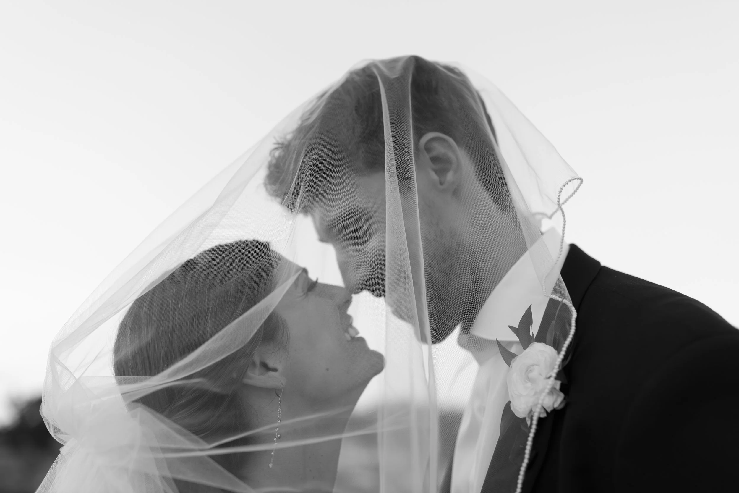 A black and white photo of a bride and groom under a veil, smiling and looking into each other's eyes.