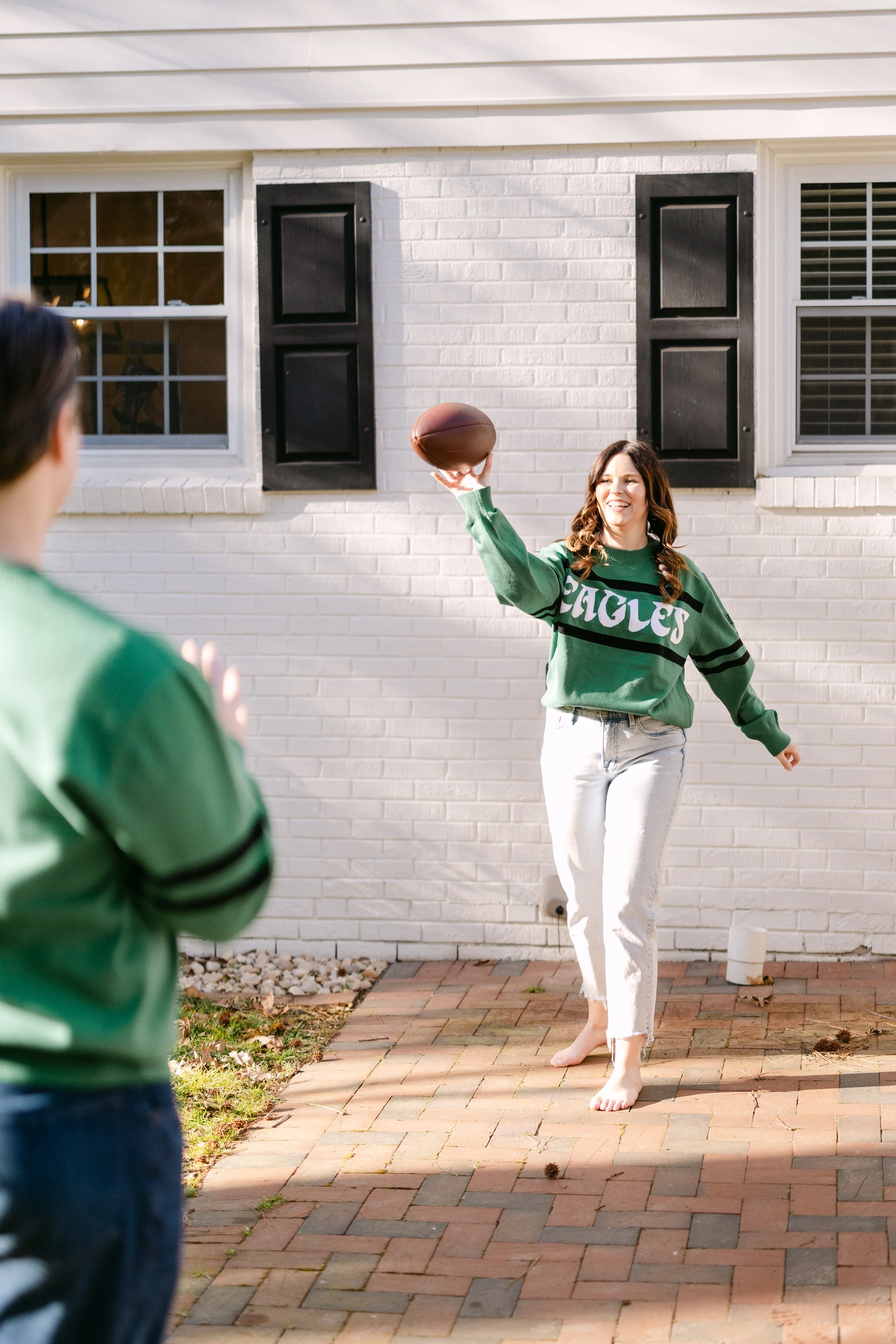A woman in a green sweatshirt and white jeans playing football with a child outdoors on a brick patio in front of a white brick house.