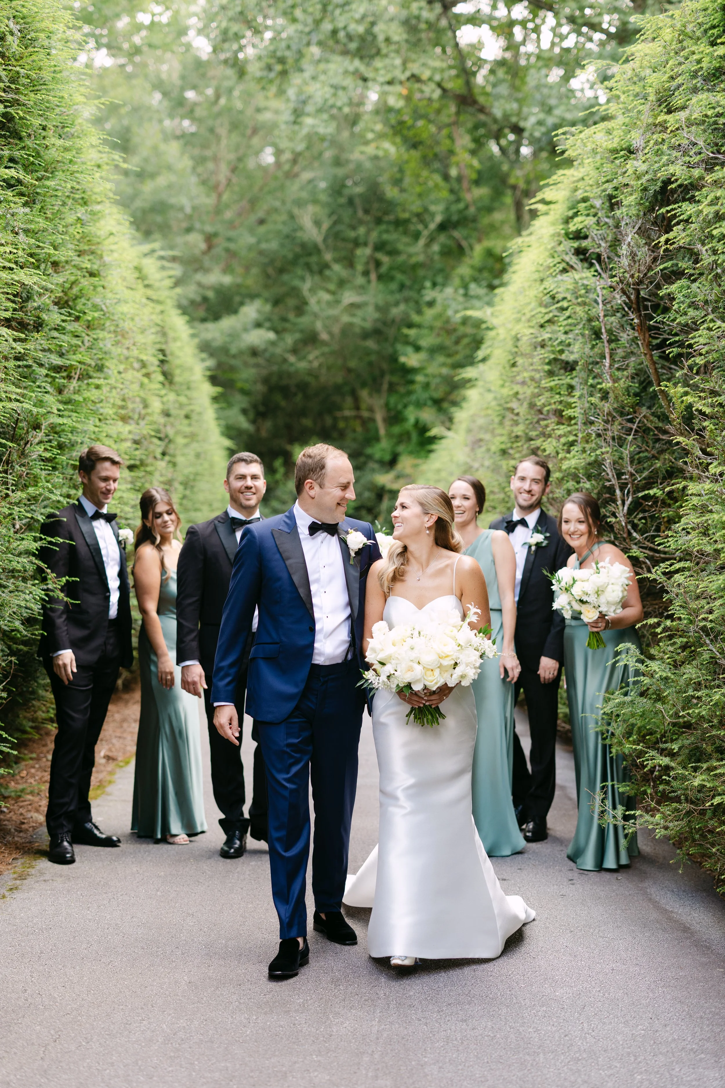 A bride and groom smiling at each other with their wedding party in a lush green outdoor setting.