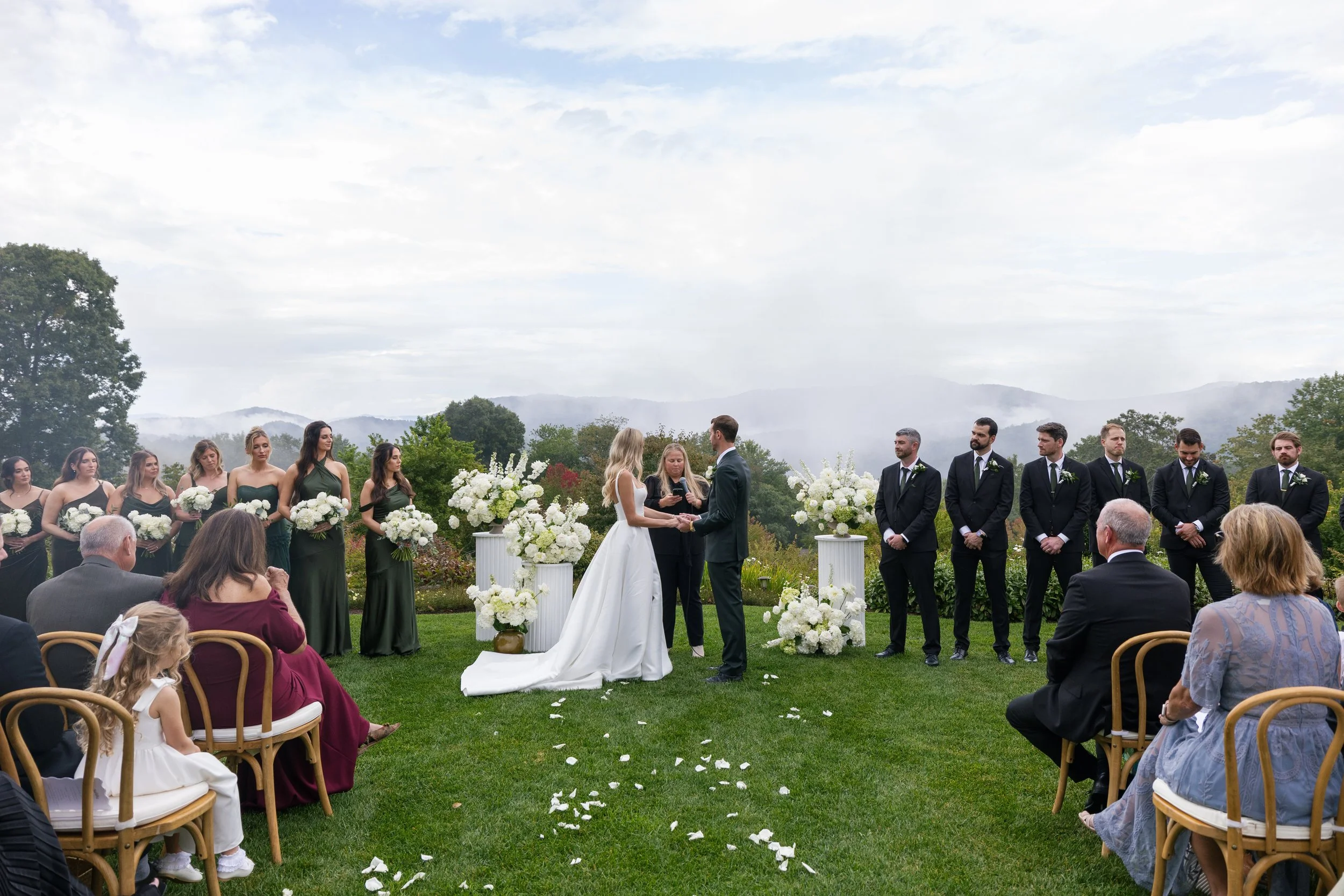 Outdoor wedding ceremony with a bride and groom exchanging vows, surrounded by bridesmaids and groomsmen, with guests seated on chairs, floral arrangements, and a scenic mountain landscape in the background.