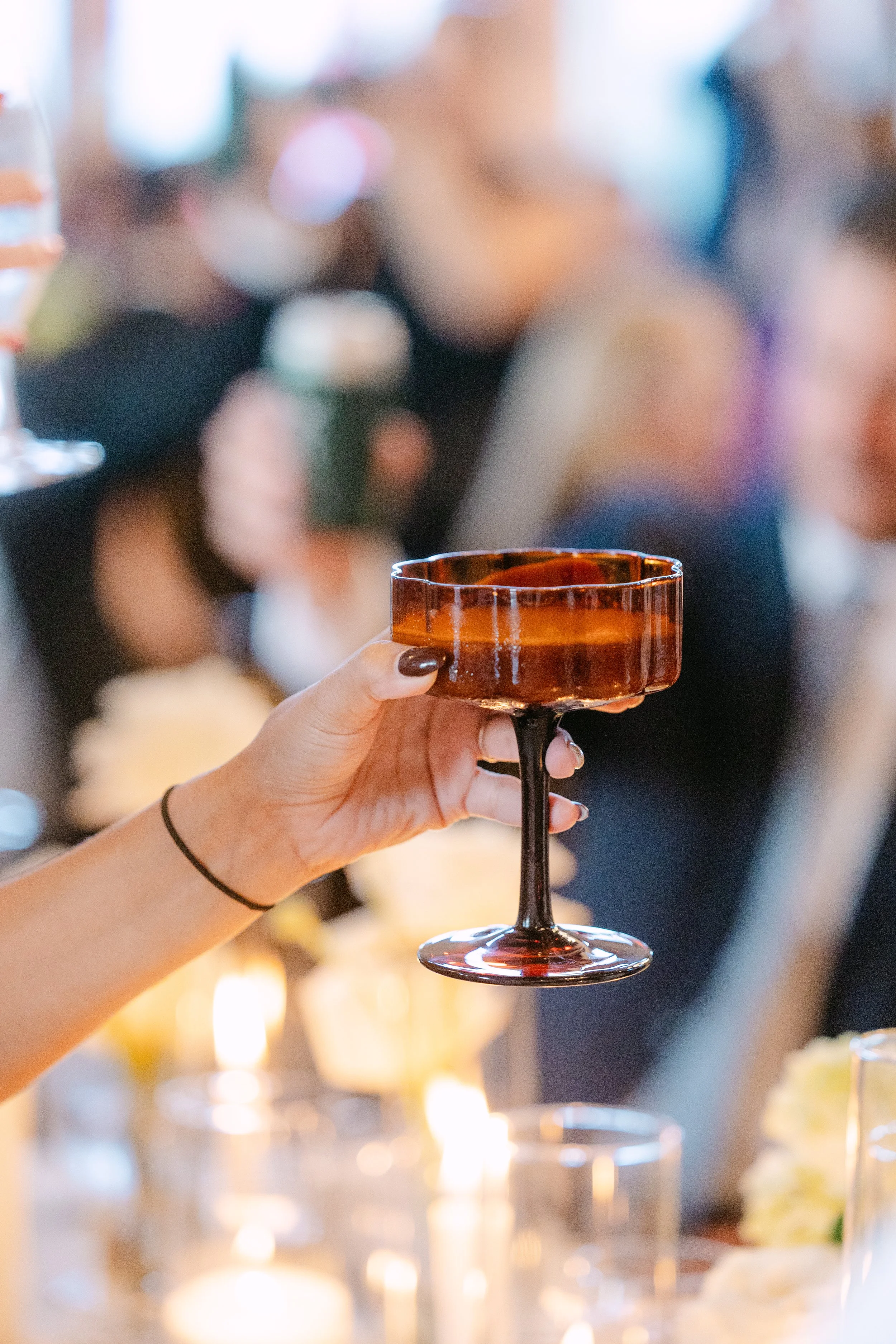 Person holding a brown cocktail glass at a social gathering with blurred people and candlelit table in the background.