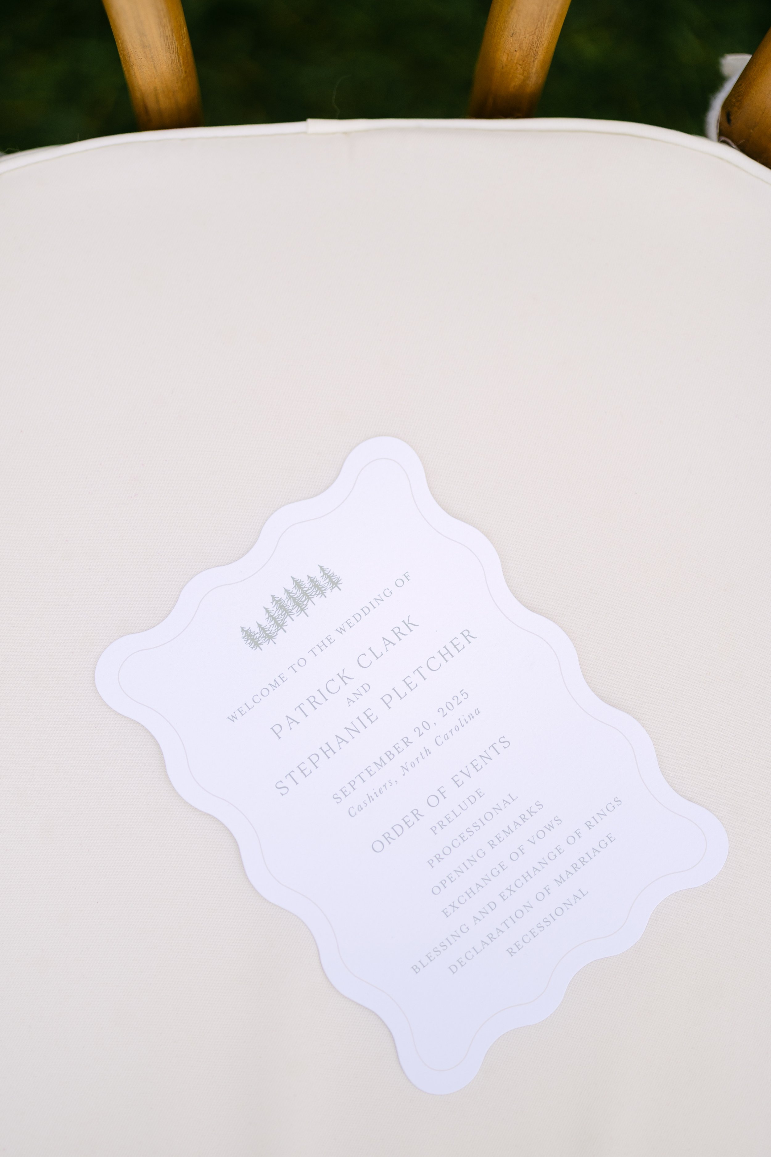 Wedding program placed on a white surface, with a background of wooden chairs and green grass.