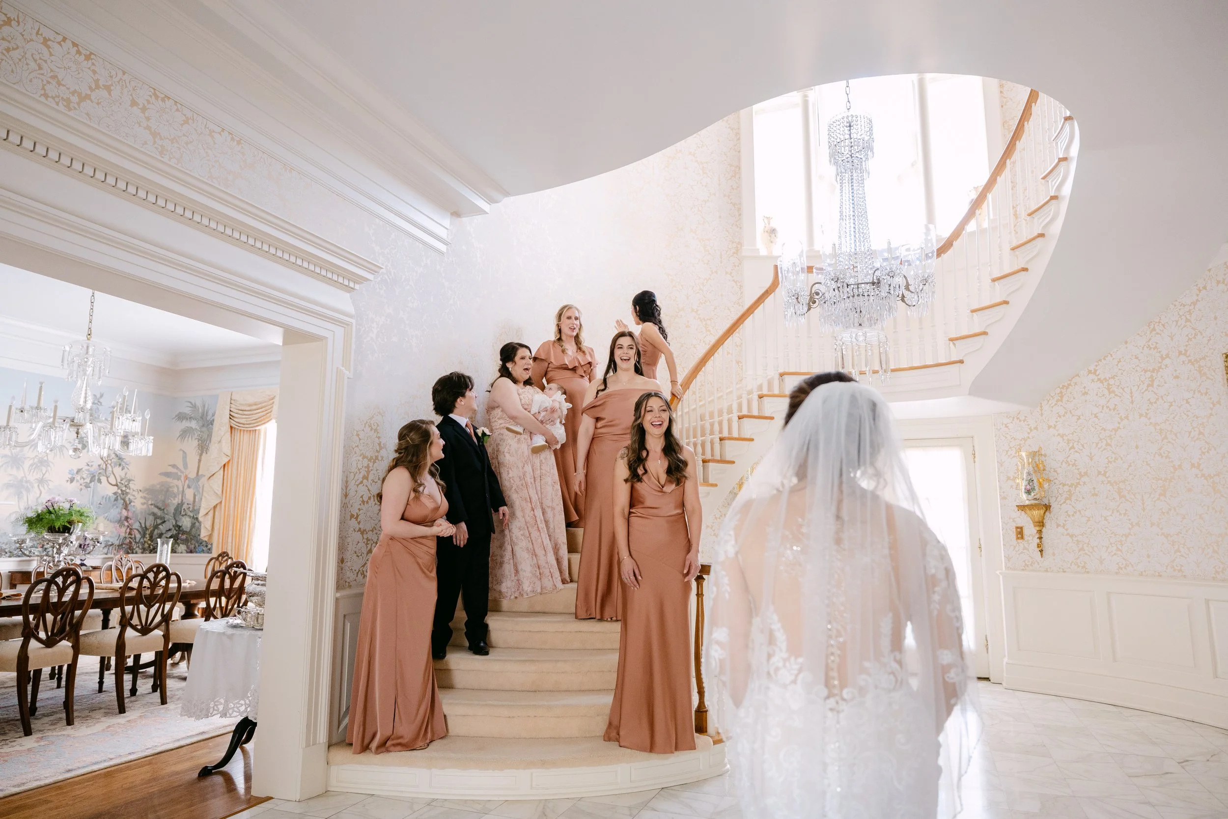 A bride in a white wedding dress with a veil stands in the foreground, facing a group of women and one man on a staircase in an elegant, well-lit interior. The women are in blush-colored bridesmaid dresses, and the man is in a dark suit. The setting 