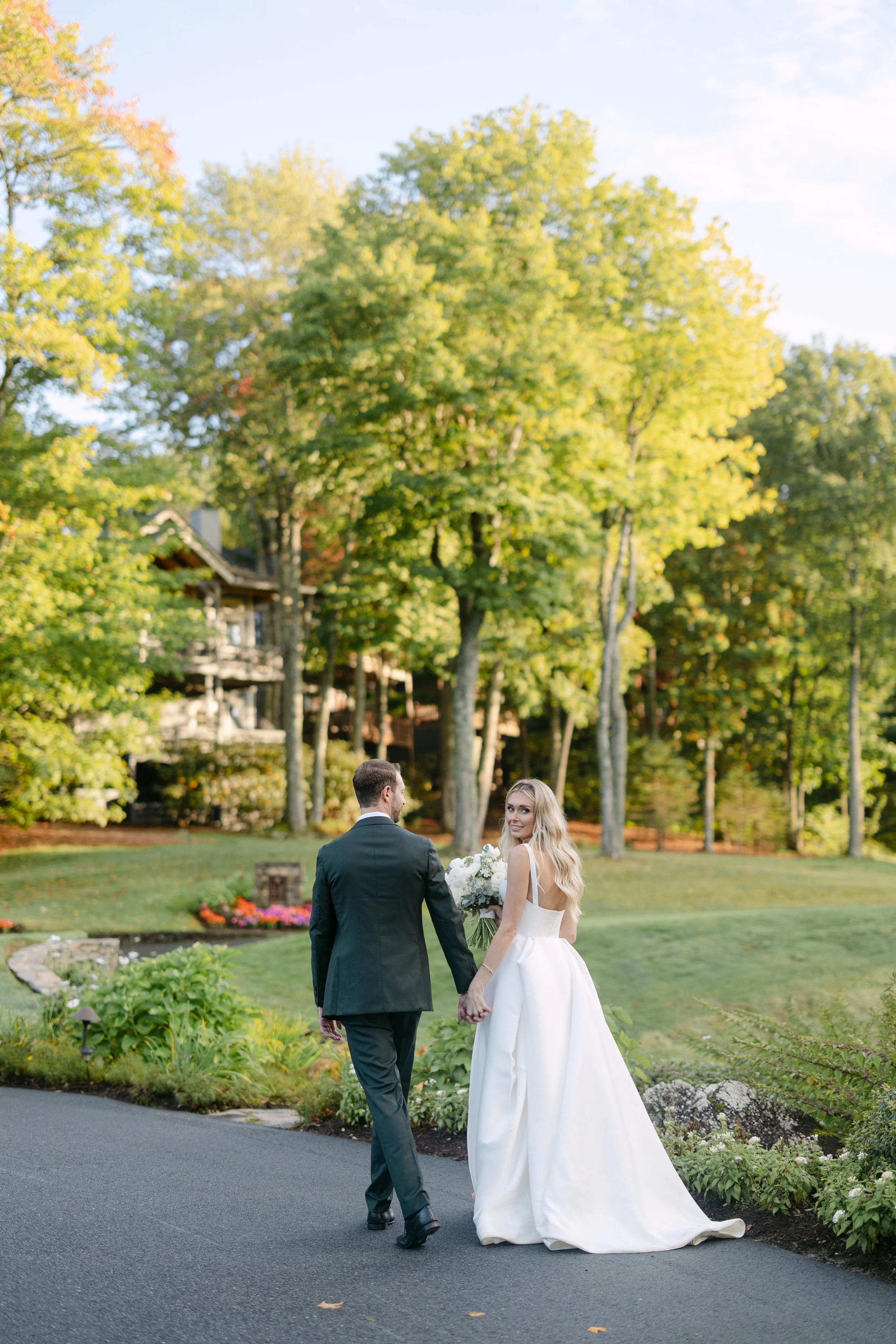 Bride and groom holding hands and walking outdoors in a garden, with the bride holding a bouquet of flowers, surrounded by trees and greenery.