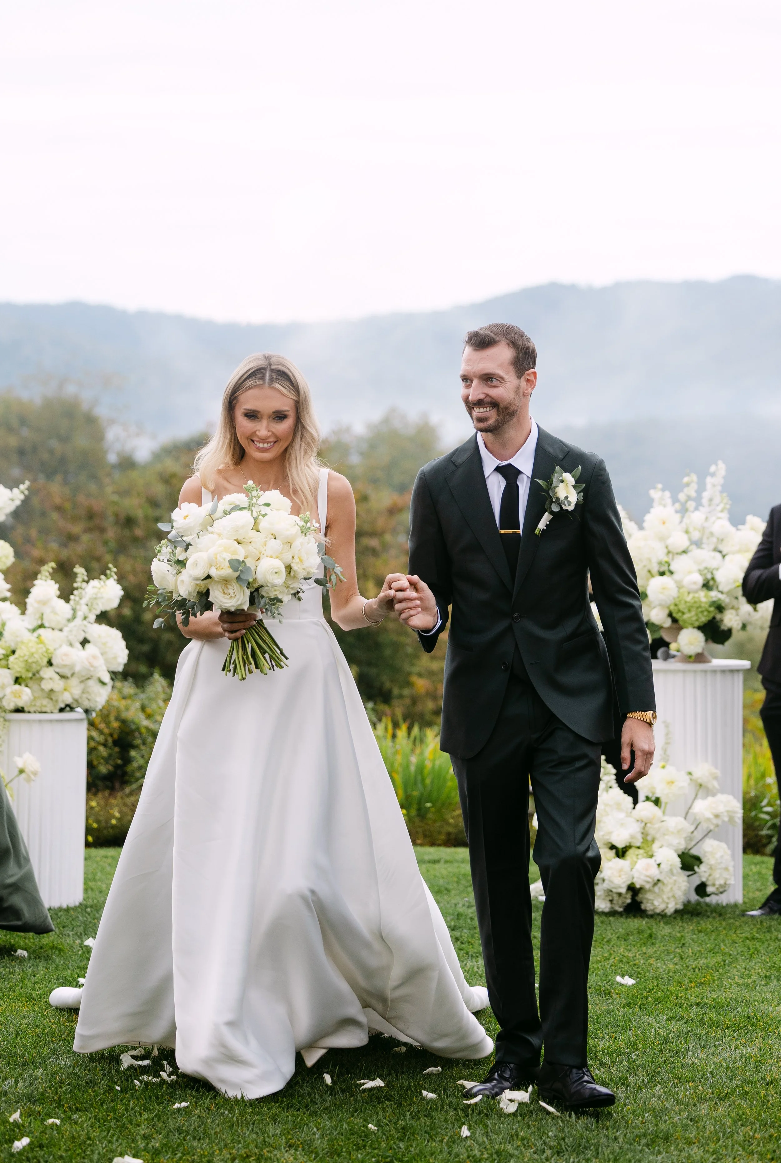 A bride and groom walking hand in hand outdoors during a wedding ceremony, surrounded by white floral arrangements, with mountains in the background.