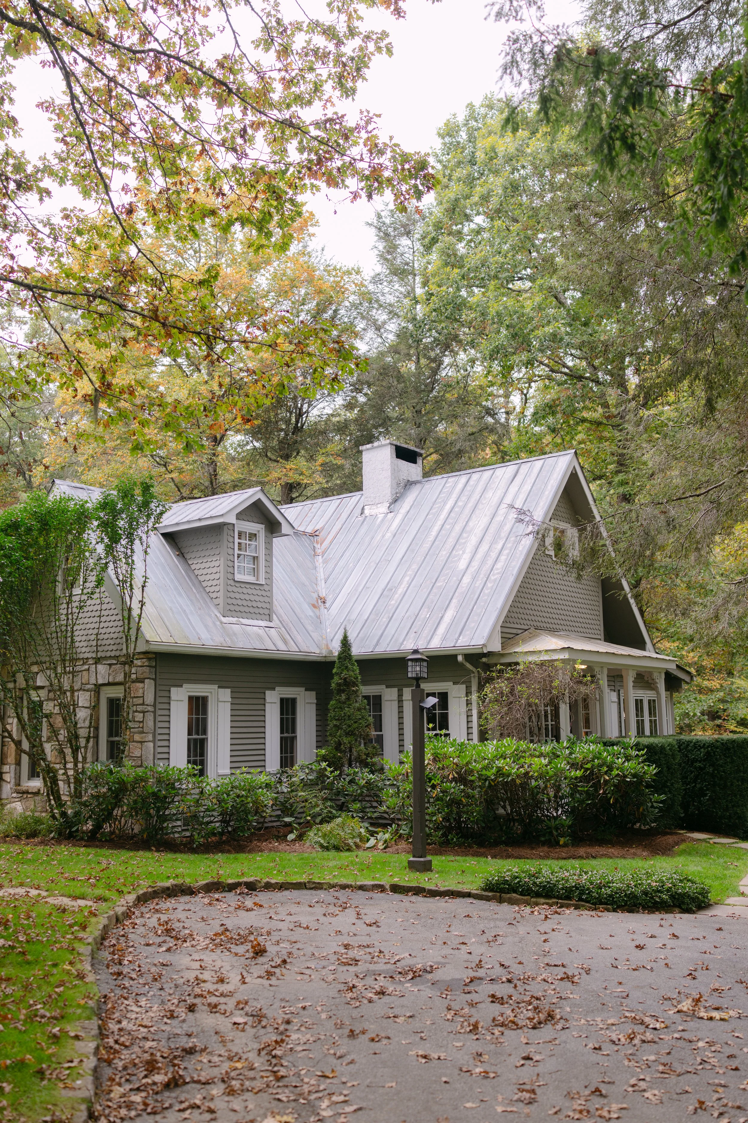 Gray cottage with a metal roof and chimney surrounded by autumn trees and landscaped shrubs