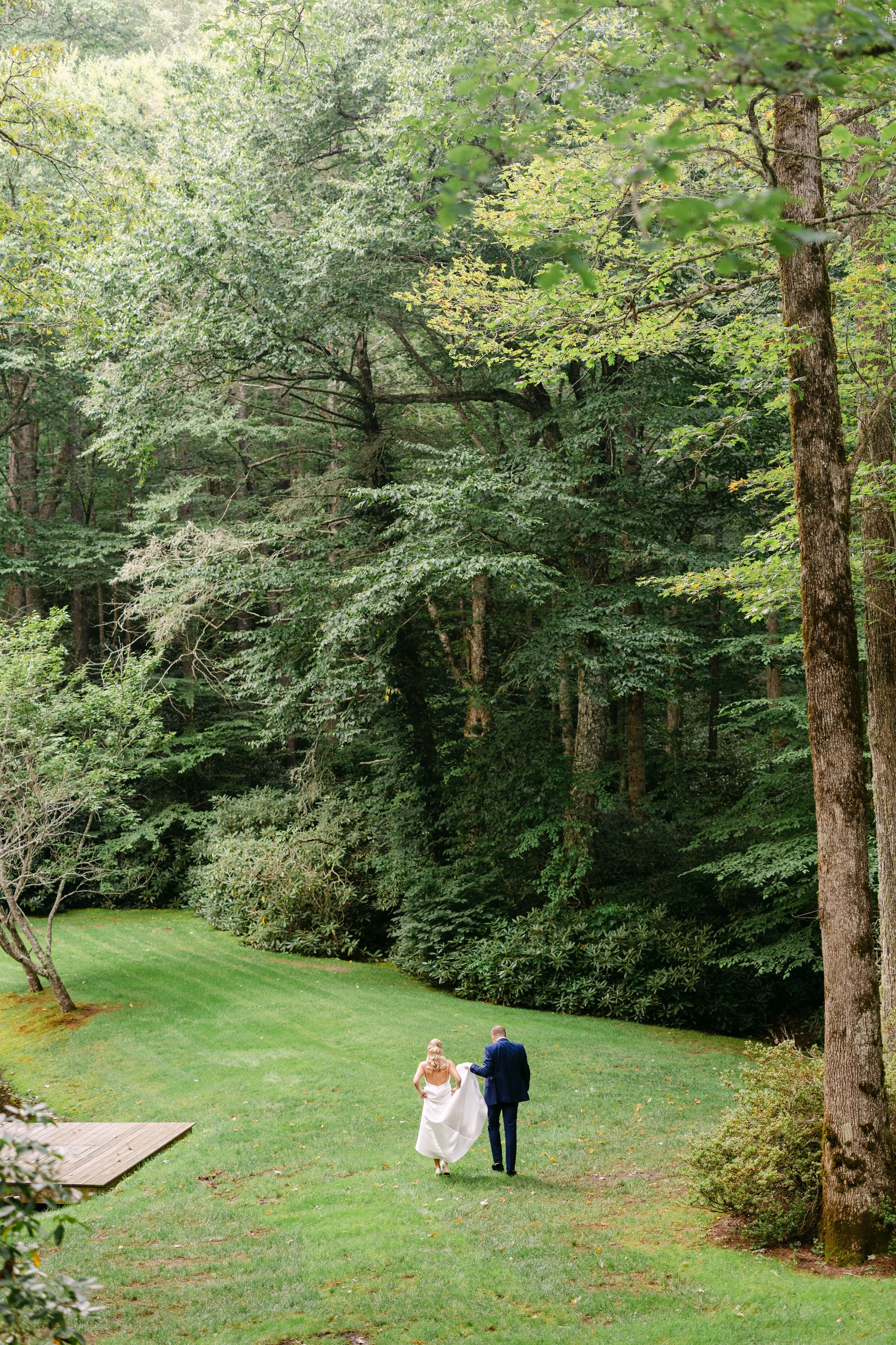 A bride and groom holding hands and walking on a grassy area in a lush, green forest setting.