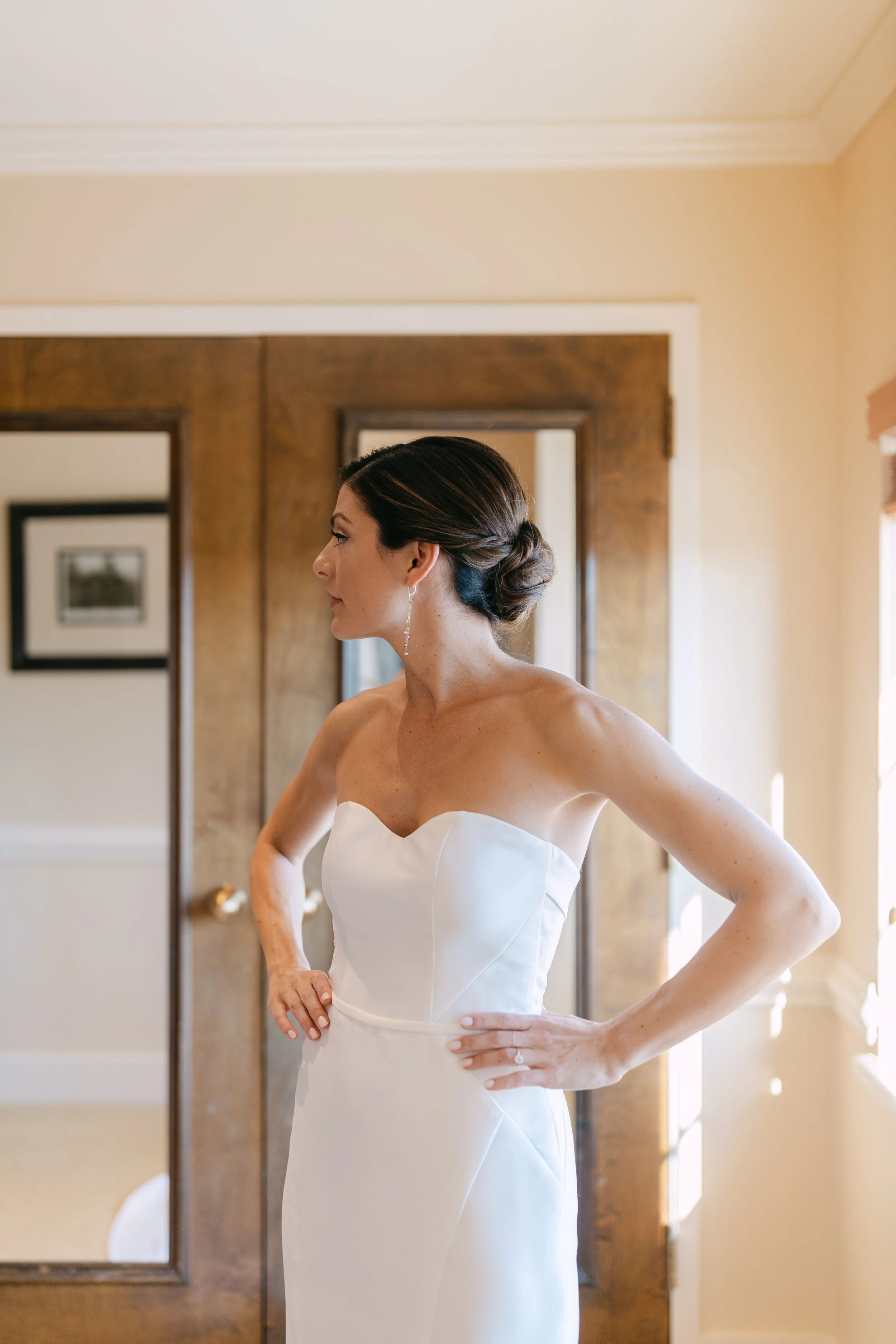A bride in a strapless white wedding dress standing indoors with her hands on her hips.