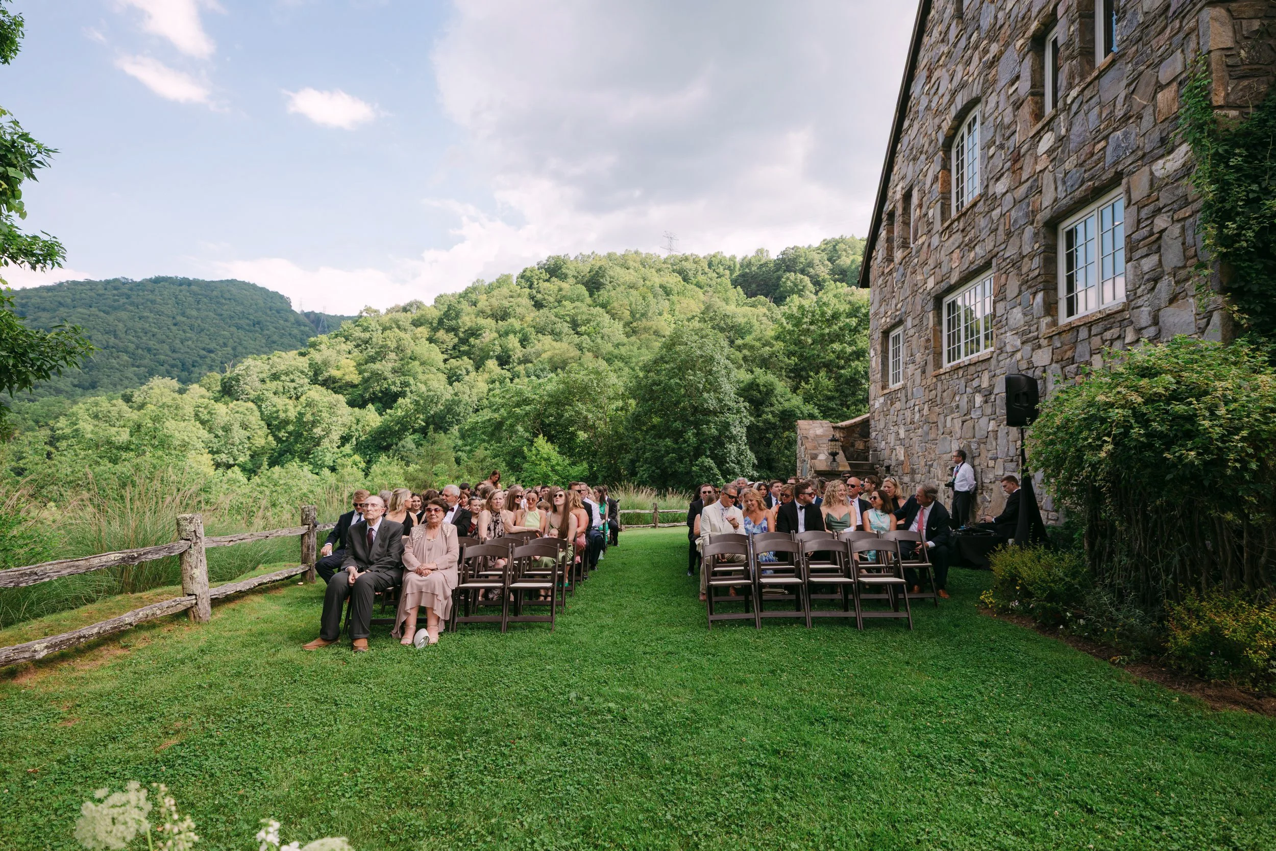 People seated outdoors on chairs at a formal event, with a stone building and green hills in the background.