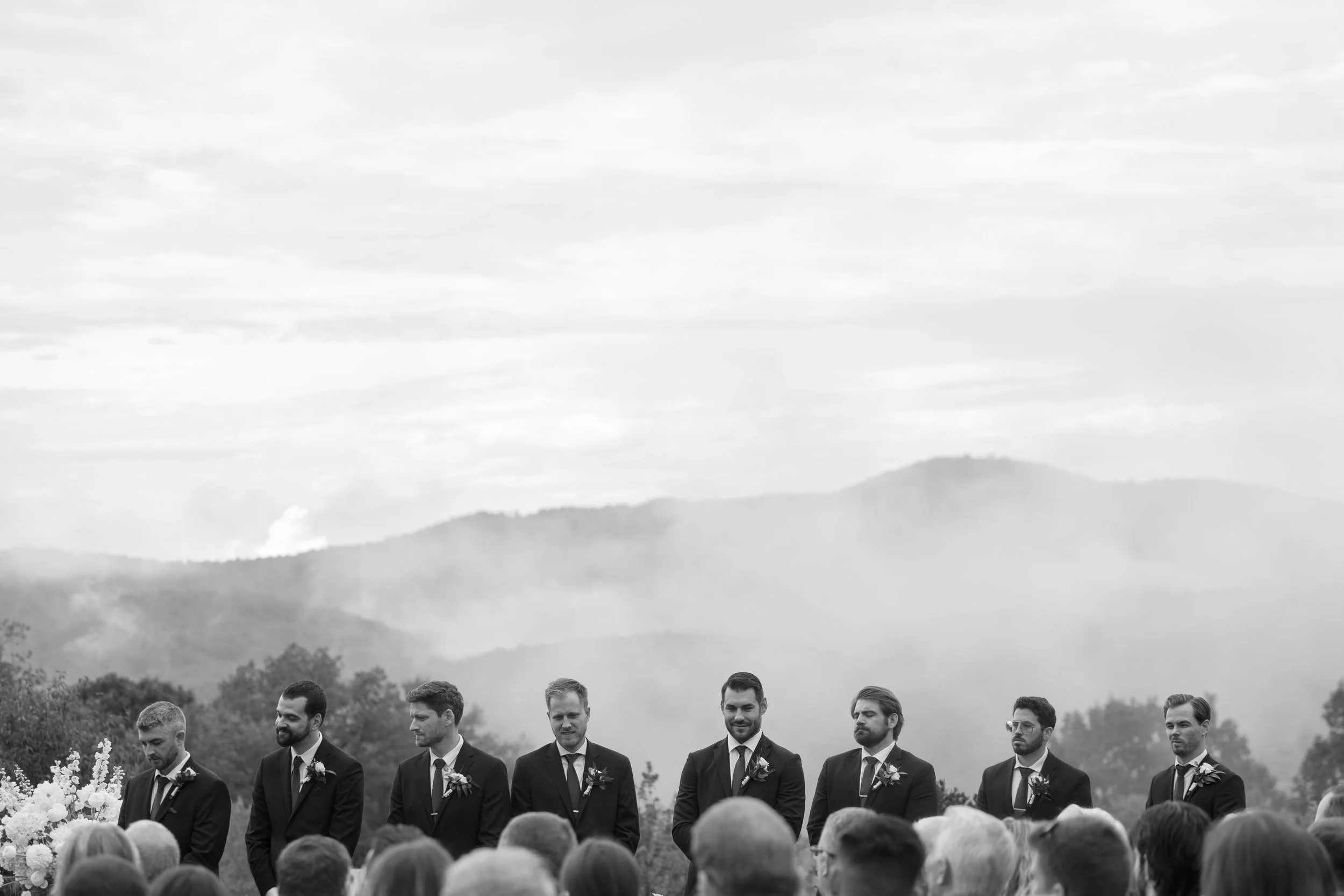 A black-and-white outdoor wedding ceremony with eight men in suits standing in front of a natural landscape with mountains and trees.
