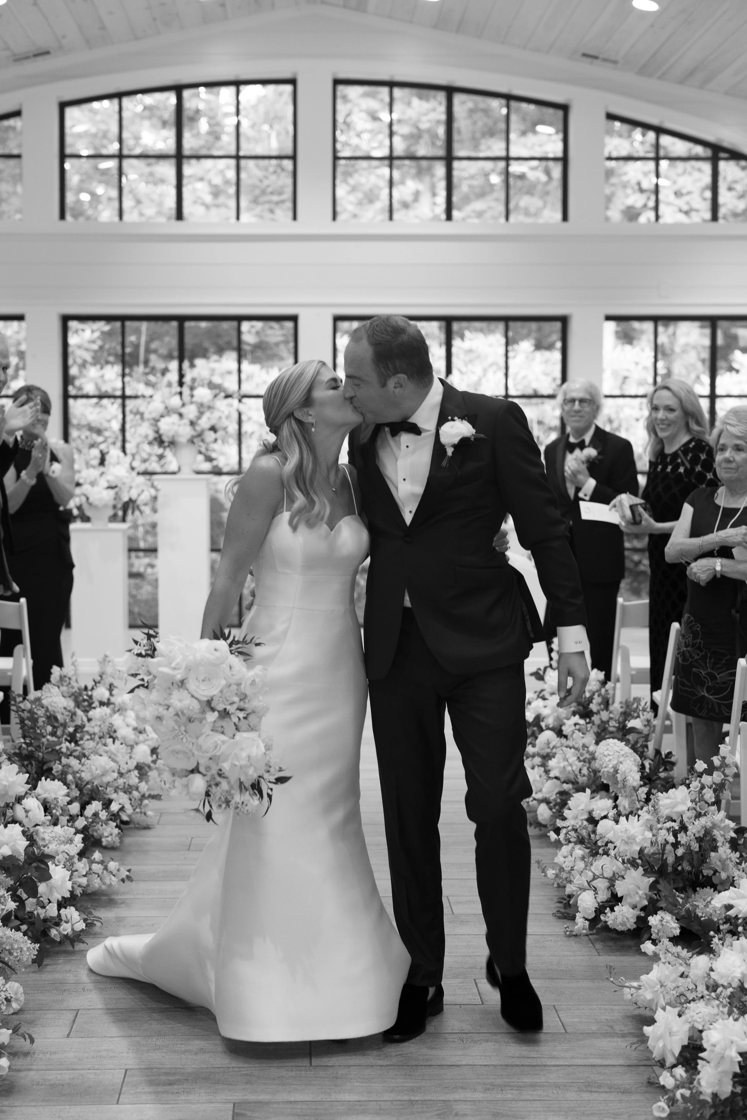A black-and-white photograph of a bride and groom kissing during their wedding ceremony indoors, surrounded by floral decorations and guests.