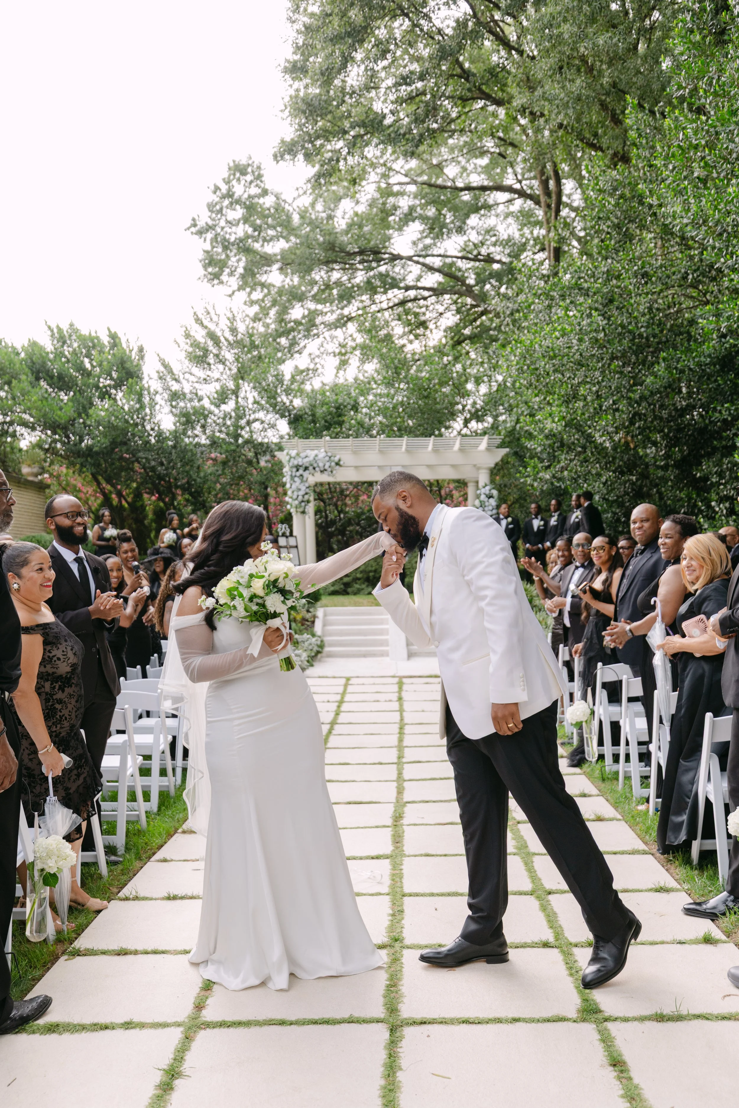 A wedding ceremony outdoors with a bride and groom at the altar, surrounded by guests. The bride is in a white dress holding a bouquet, and the groom is in a white tuxedo jacket, bow tie, and black trousers. The guests are dressed formally, smiling, 