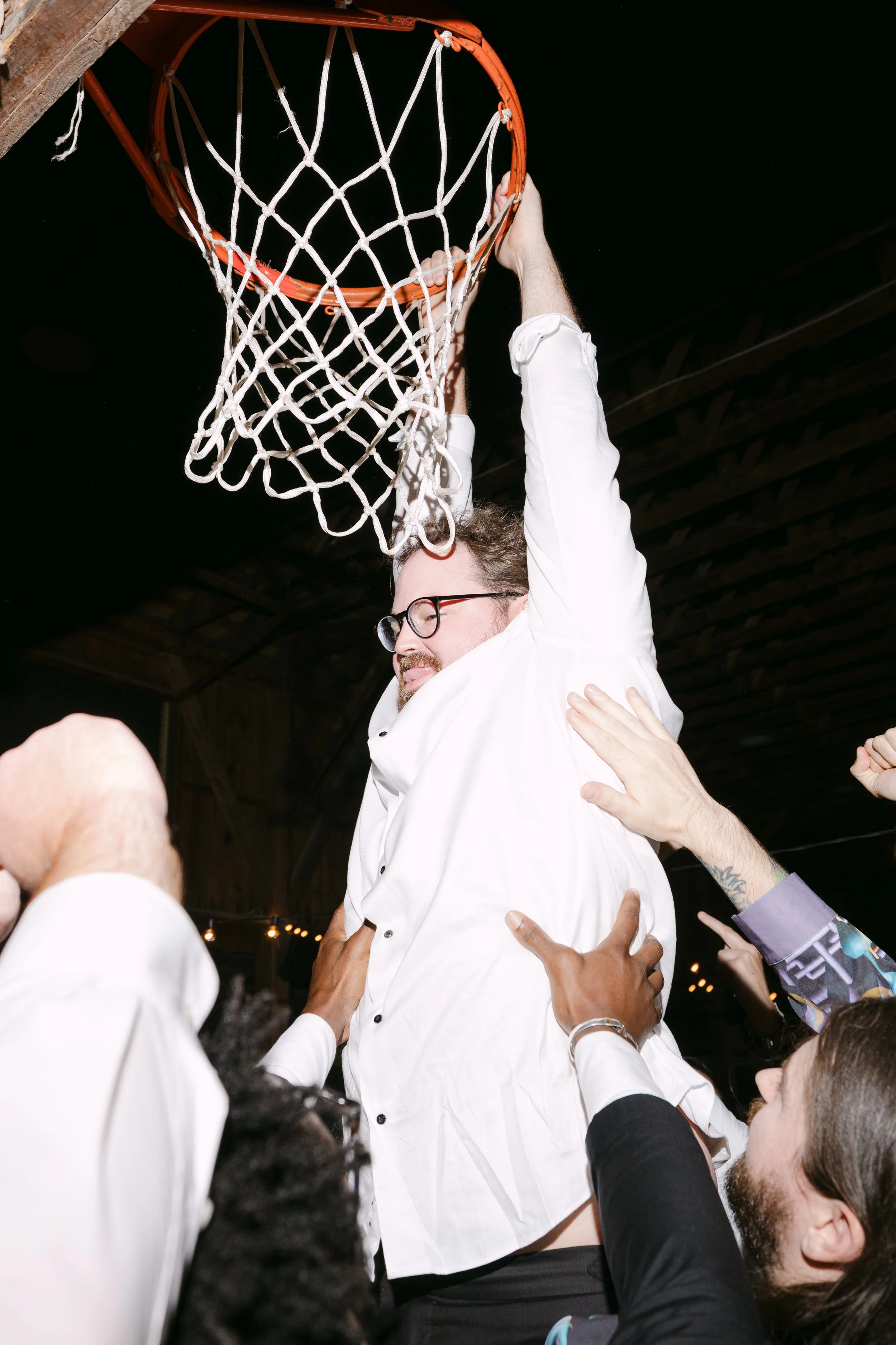 A man with glasses and curly hair, dressed in a white shirt, is being lifted and celebrated by others around a basketball hoop. The scene appears to be a joyful moment during a celebration or party.