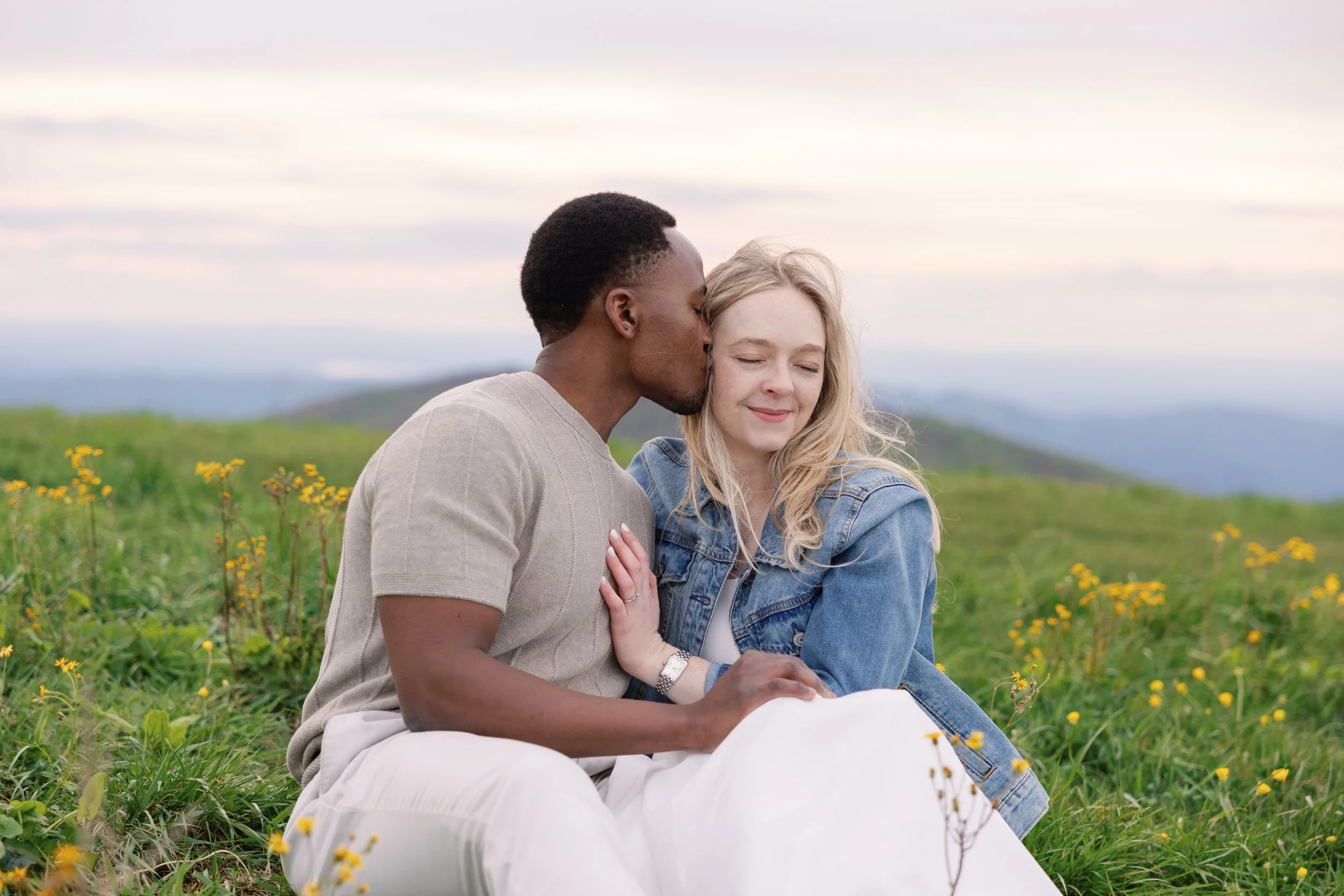 A young couple sitting in a field of yellow flowers, with the man kissing the woman's temple as she smiles with her eyes closed.