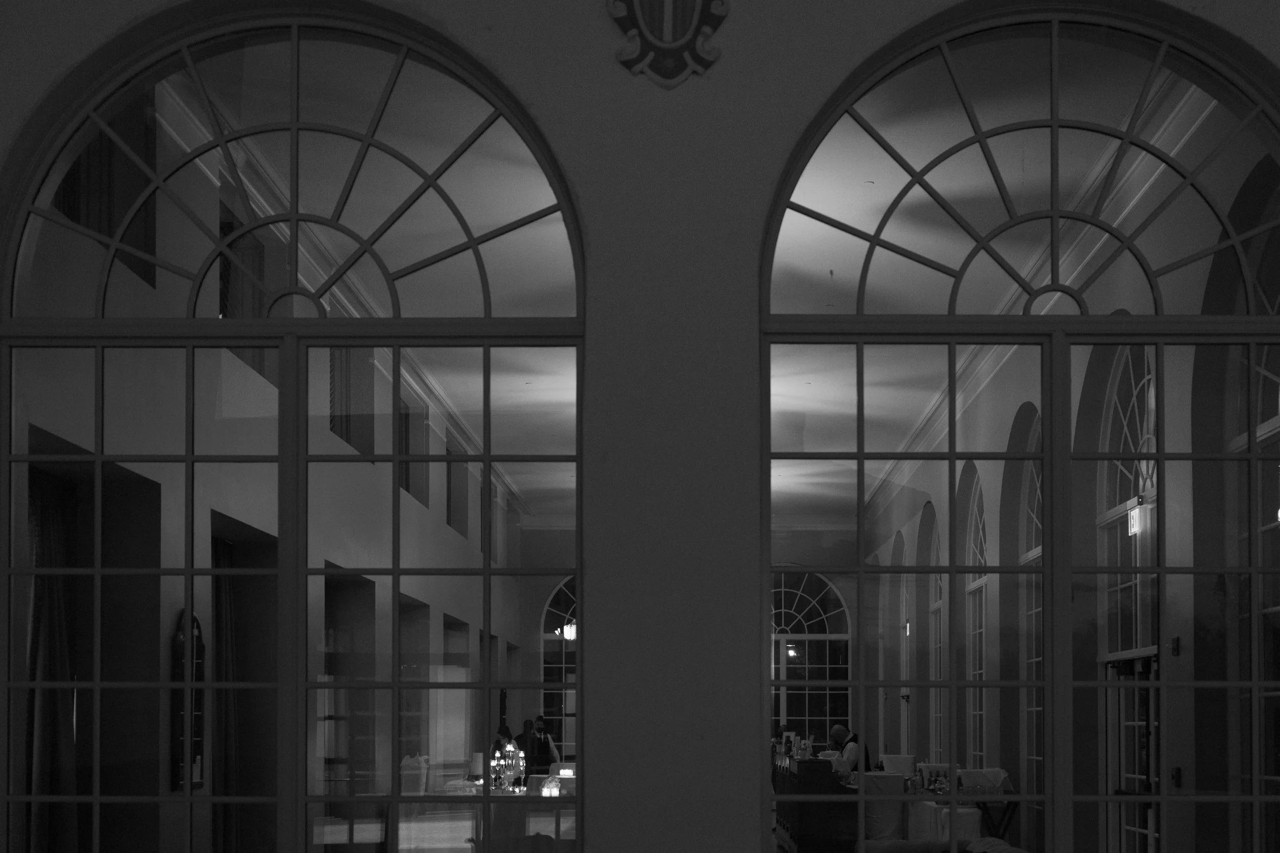 Interior of a building with arched windows, view through glass panels showing a dimly lit room with a person sitting at a table, and a representative ambiance of a formal event.
