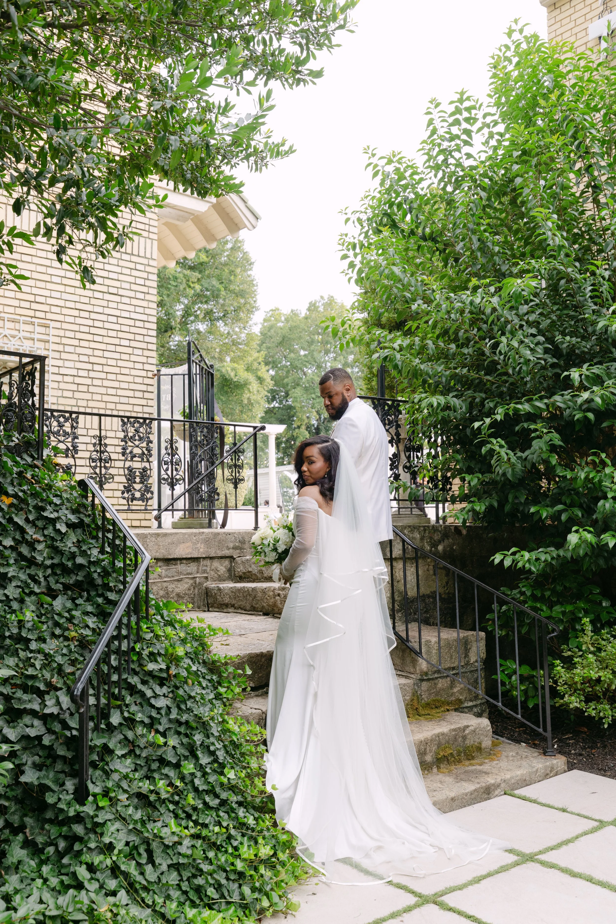 A bride and groom standing on stone steps outside a brick house, surrounded by lush green trees and bushes, during a wedding ceremony.