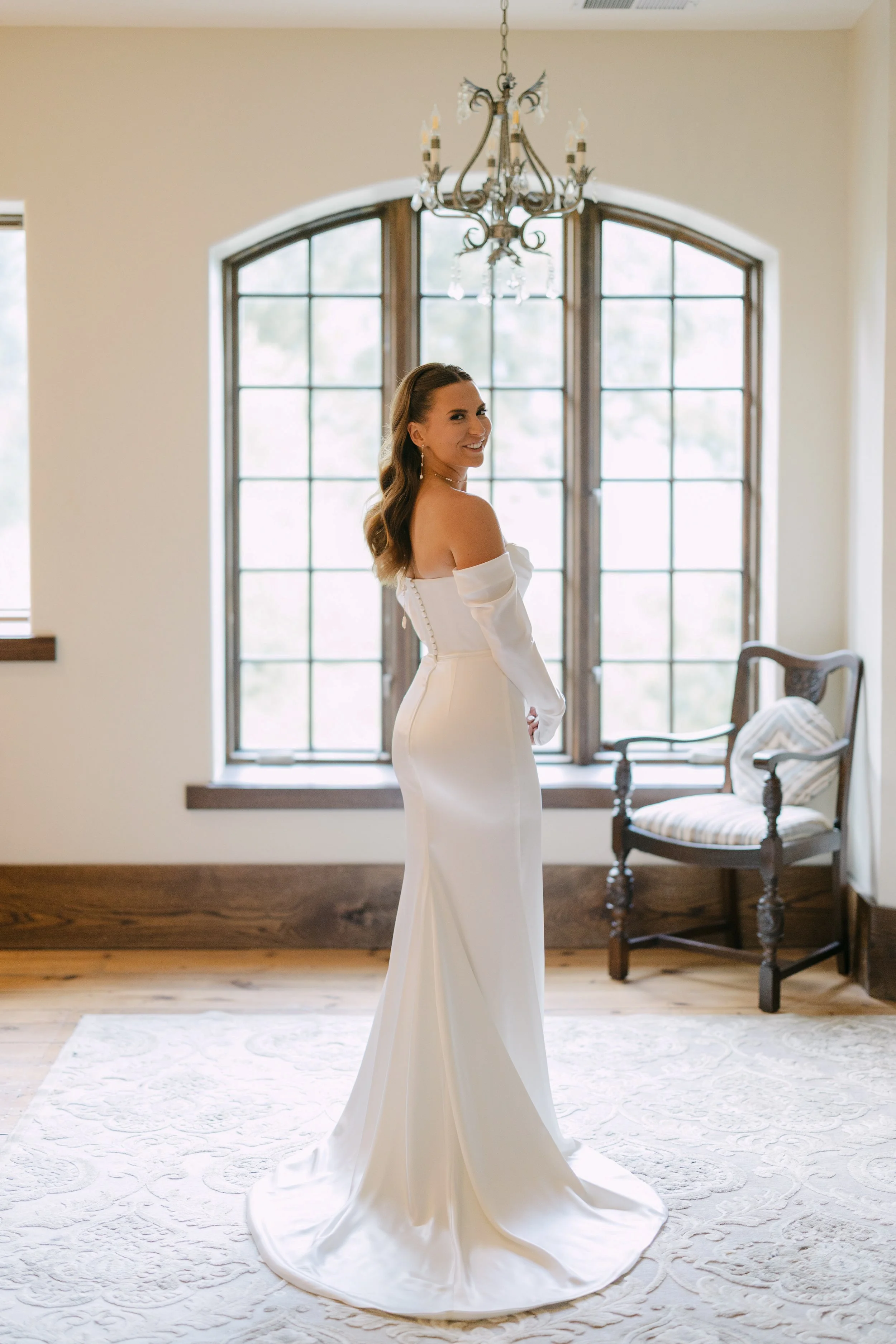 A woman in a white off-shoulder wedding dress standing indoors by a large window, smiling at the camera.