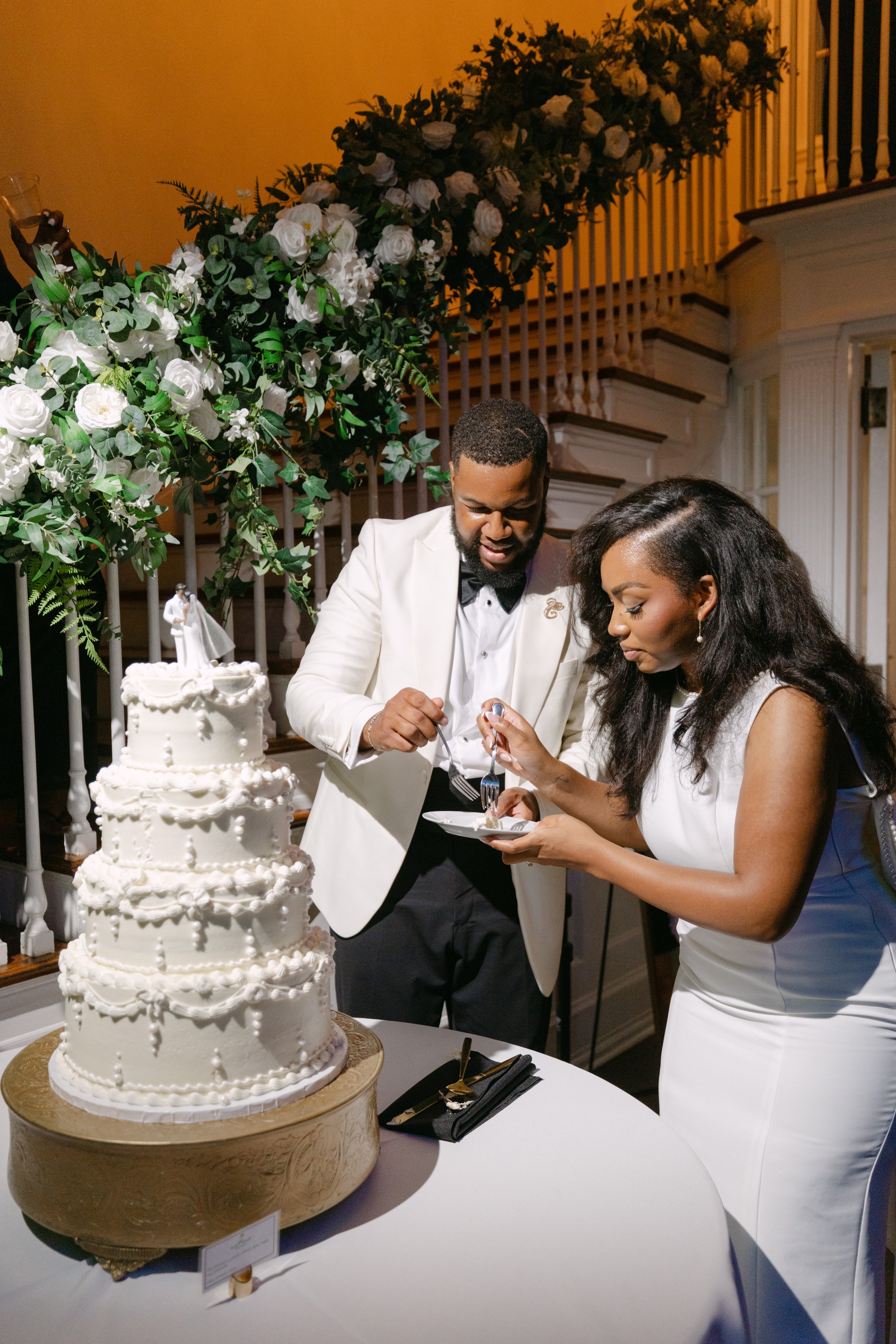 A couple dressed in formal wedding attire, the man in a white tuxedo jacket and the woman in a white dress, are cutting a multi-tiered white wedding cake with decorative piping and small sugar pearls. The scene is decorated with large green foliage a