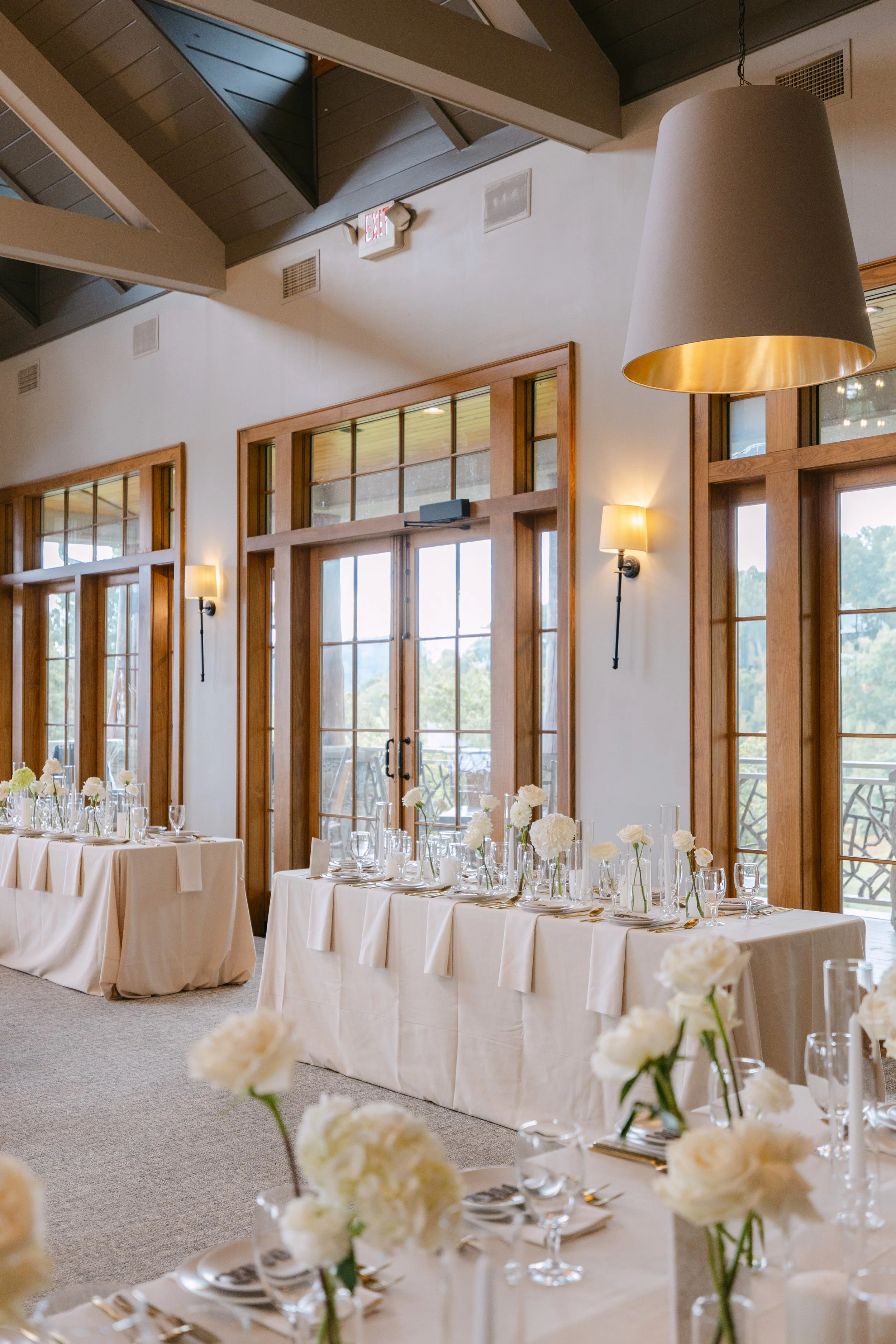 Elegant indoor event space with large windows, cream tablecloths, white flower centerpieces, and glassware arranged for a formal occasion.