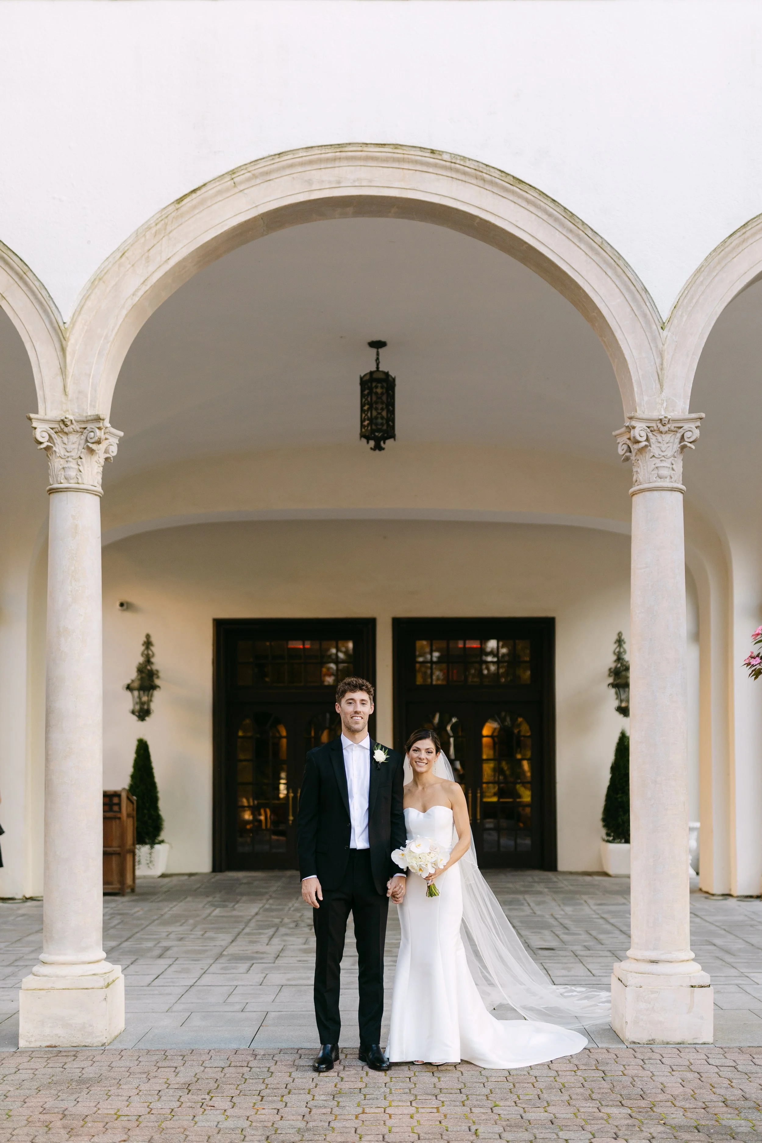 A bride and groom standing together outside a building with archways, holding hands and smiling, dressed in wedding attire with the bride holding a bouquet.