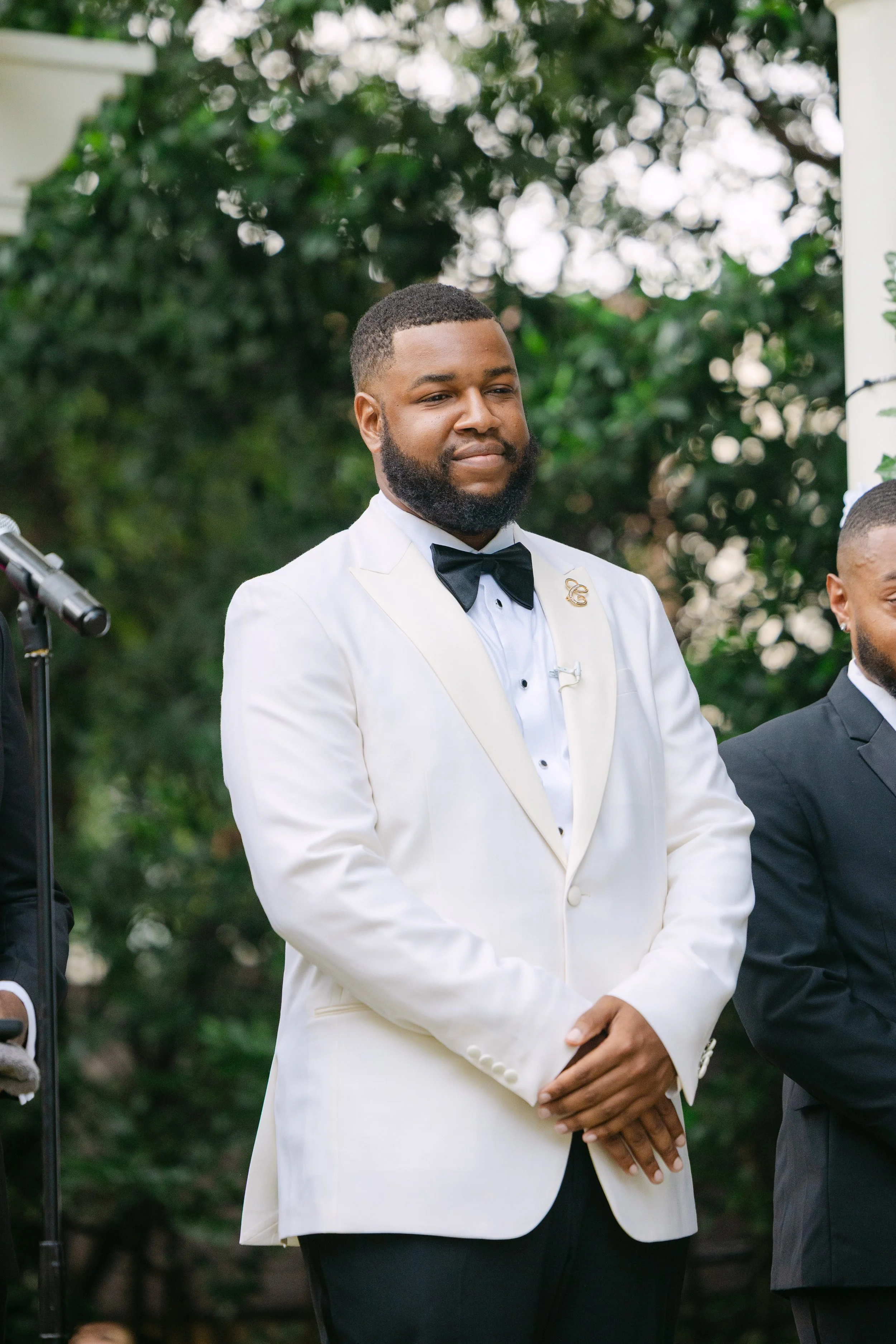 A man dressed in a white tuxedo with a black bow tie and a 'G' pin on his lapel, standing outdoors during daytime with greenery in the background, participating in a formal event or ceremony.