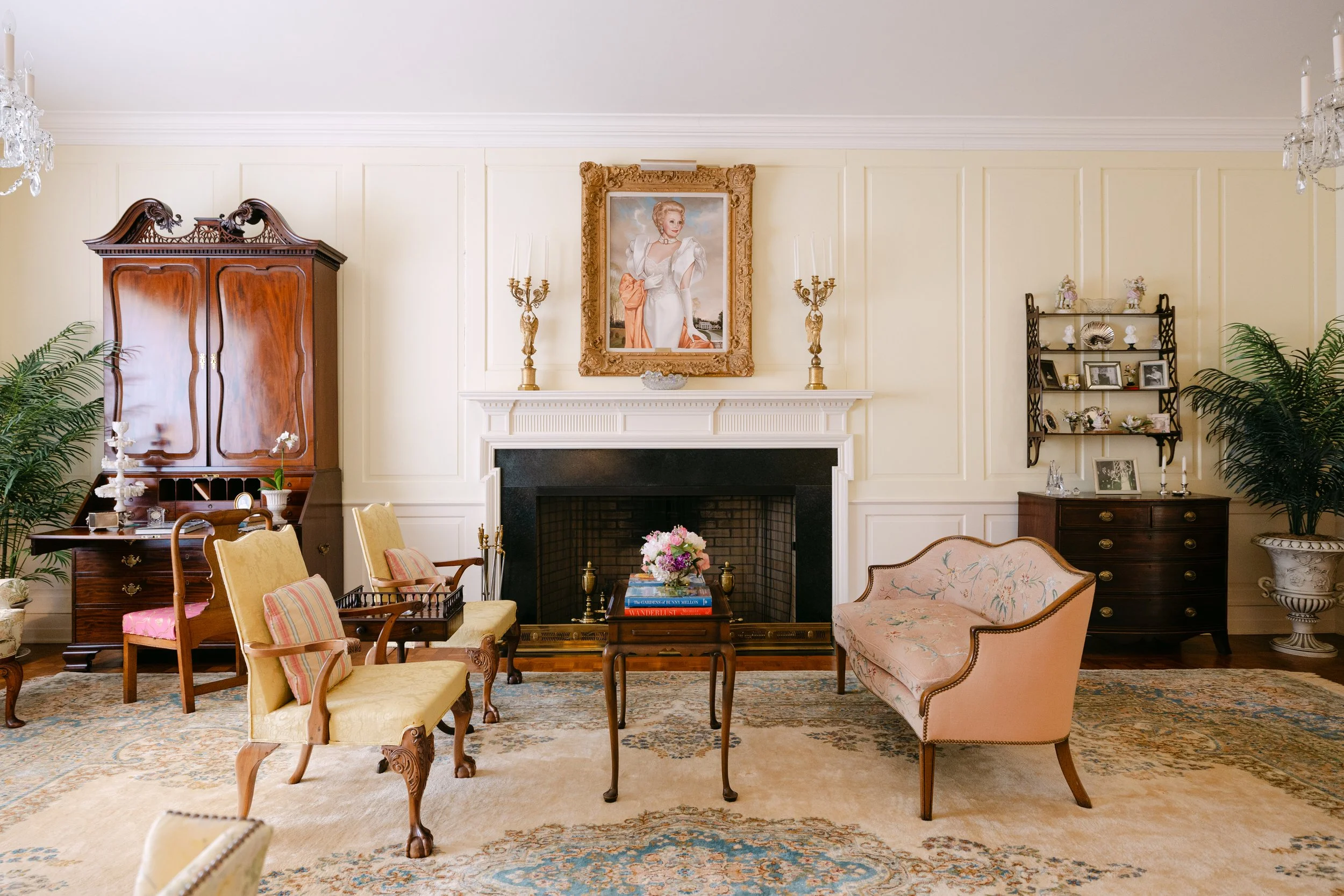 Living room with vintage furniture, a fireplace, framed portrait above the mantel, decorative plants, and shelves with ornaments and photographs.