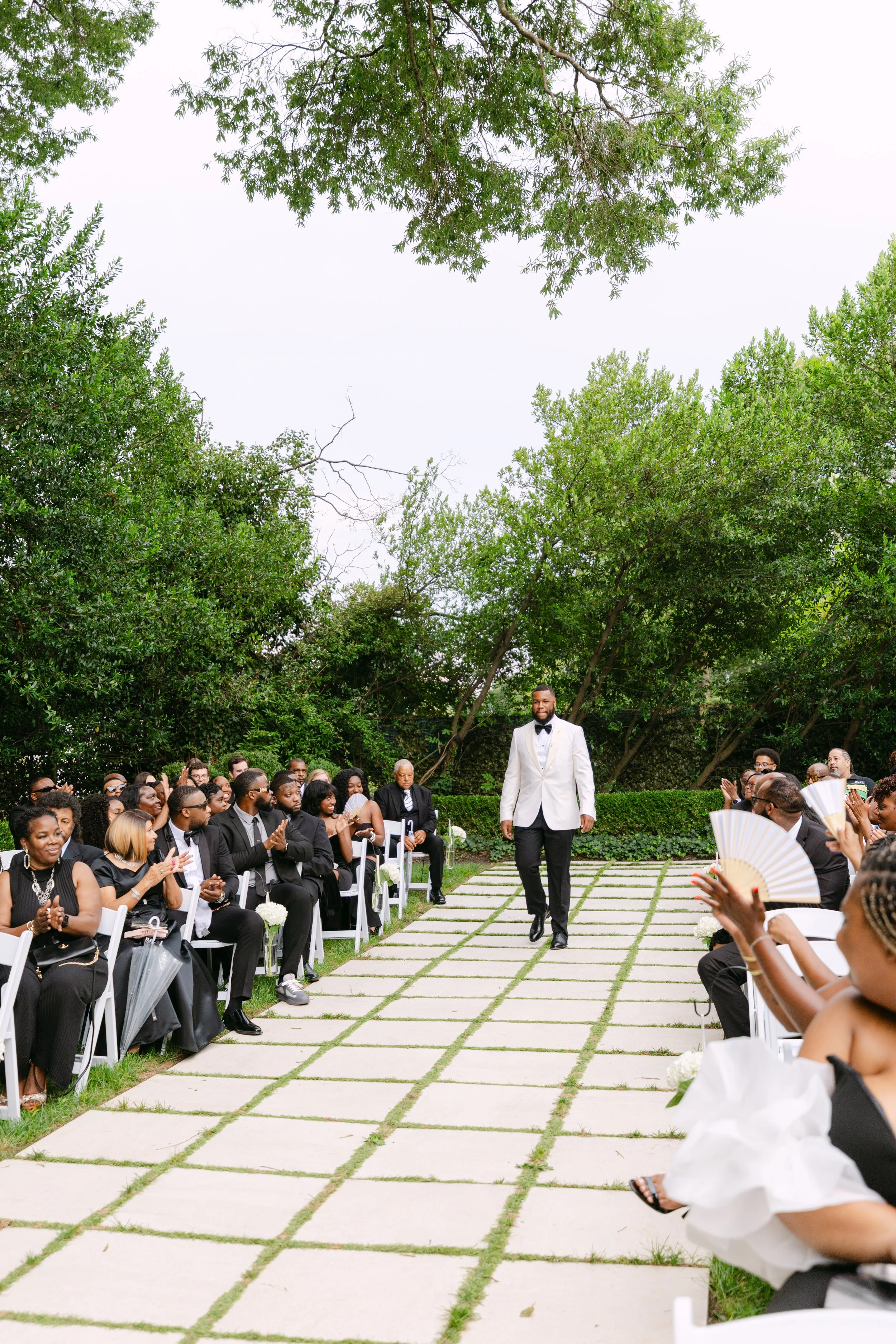 A wedding ceremony outdoors with a man in a white tuxedo walking down the aisle, surrounded by seated guests wearing black attire, under green trees.
