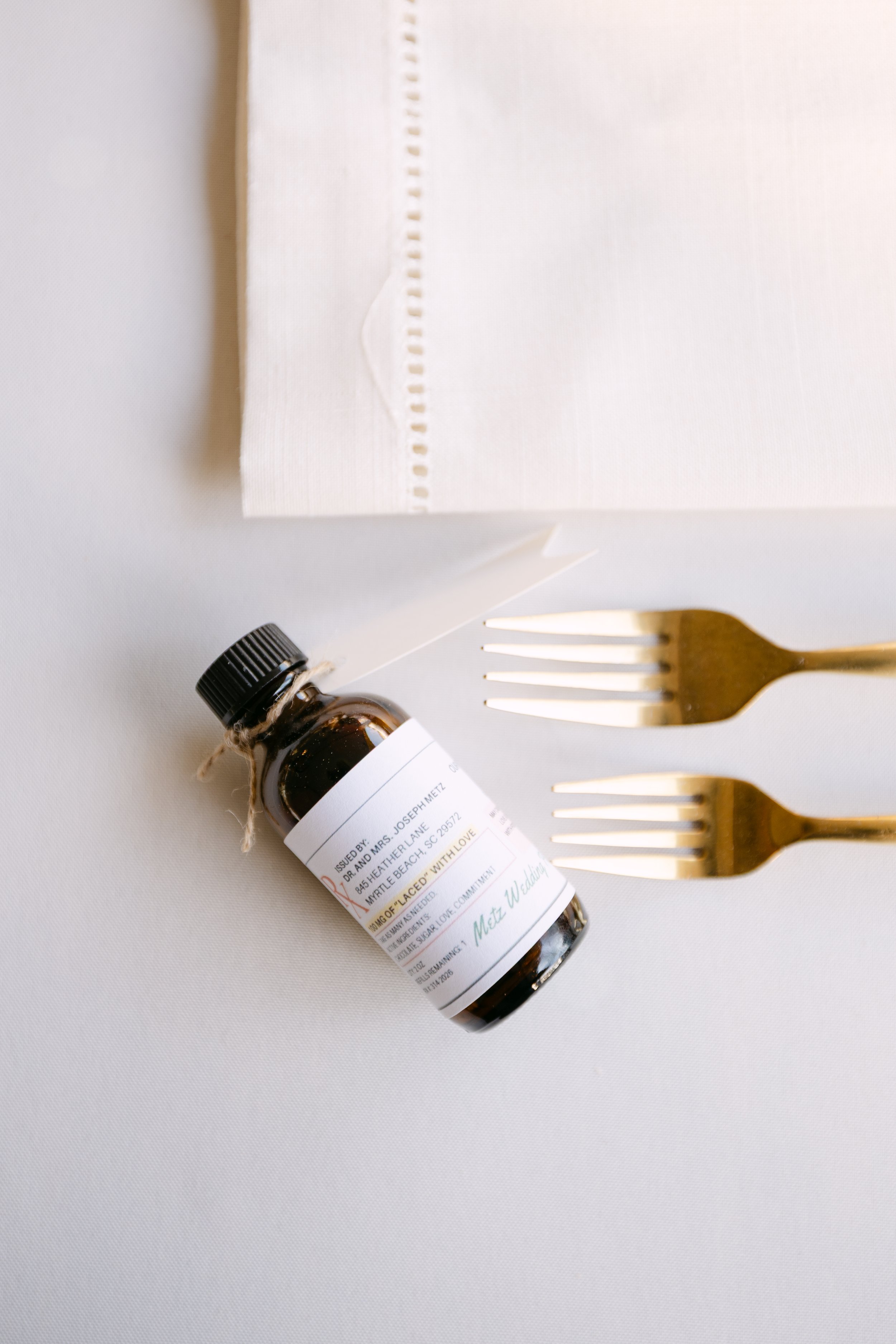 A small amber glass bottle with a black cap and a white label, placed on a white surface next to a white tissue box and two gold forks.