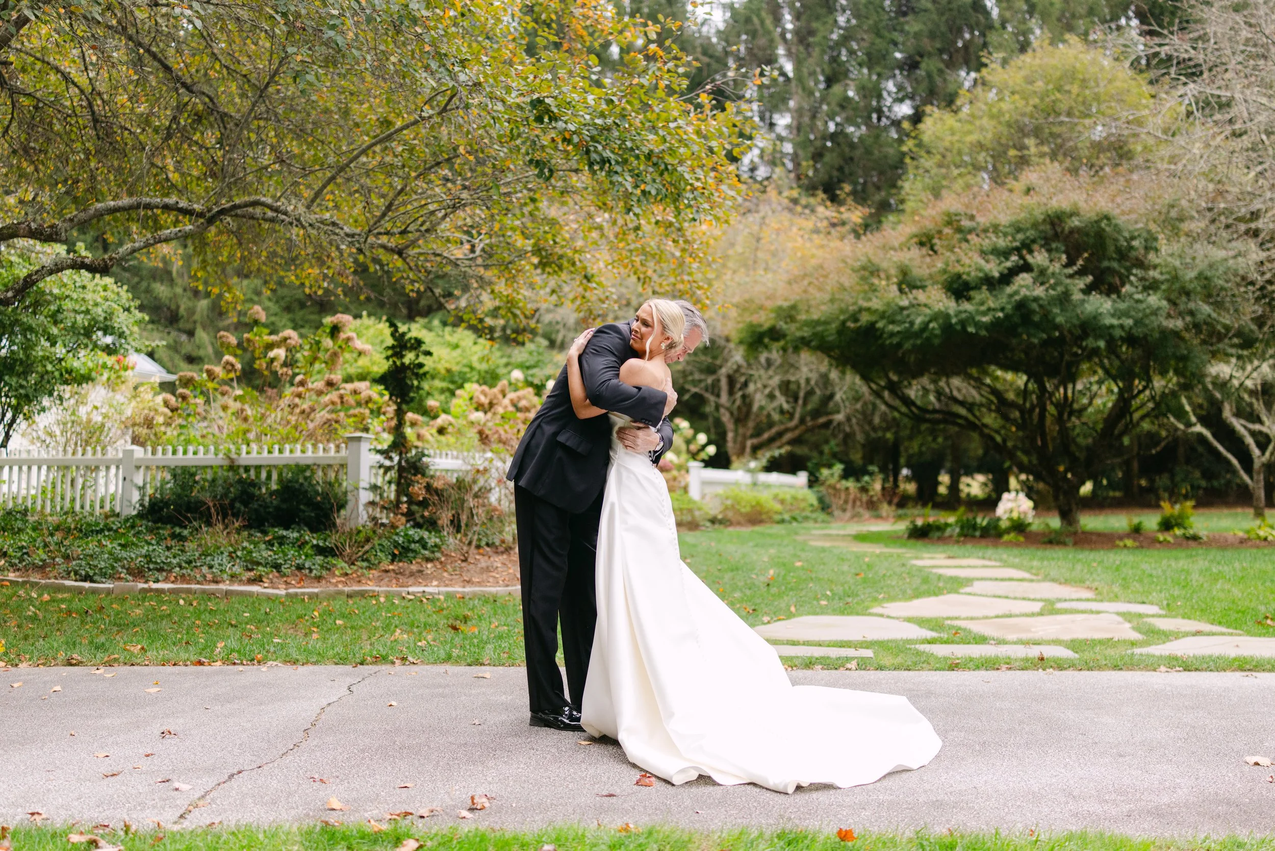 Bride and groom embracing outdoors in a garden, with the bride’s long wedding dress train spread across the pavement.