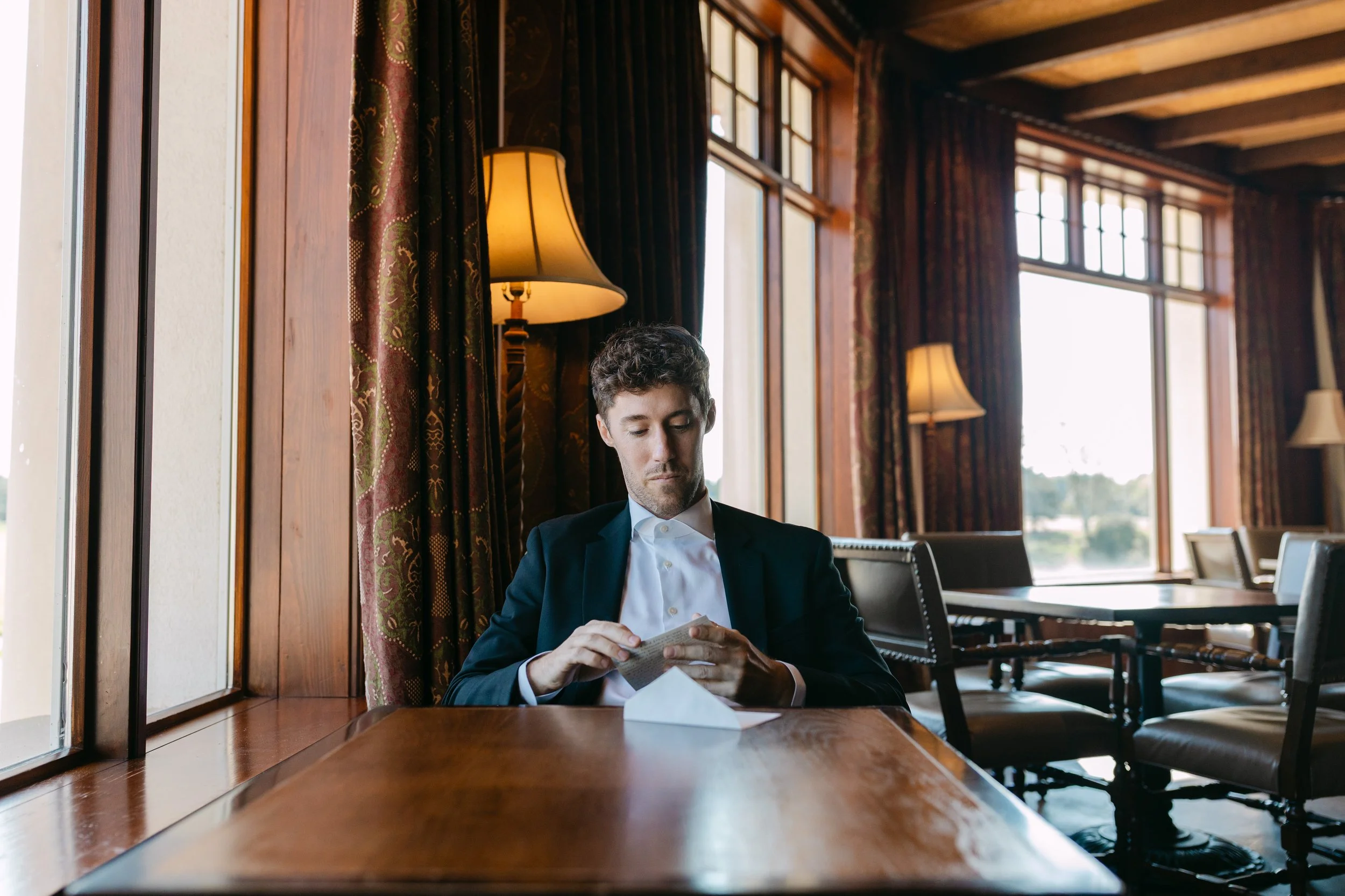 A man in a suit sitting at a wooden table in a warmly lit room with large windows, reading a small pamphlet or booklet.