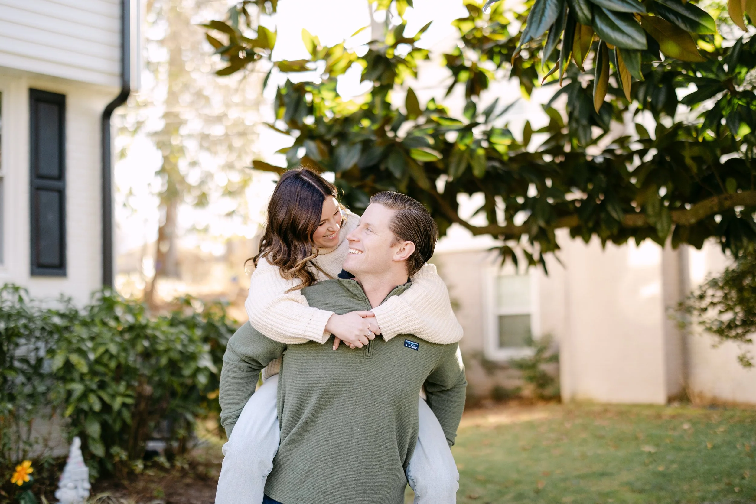 A happy couple outdoors, with the woman on the man's back, smiling and looking at each other in a garden with a large tree and house in the background.