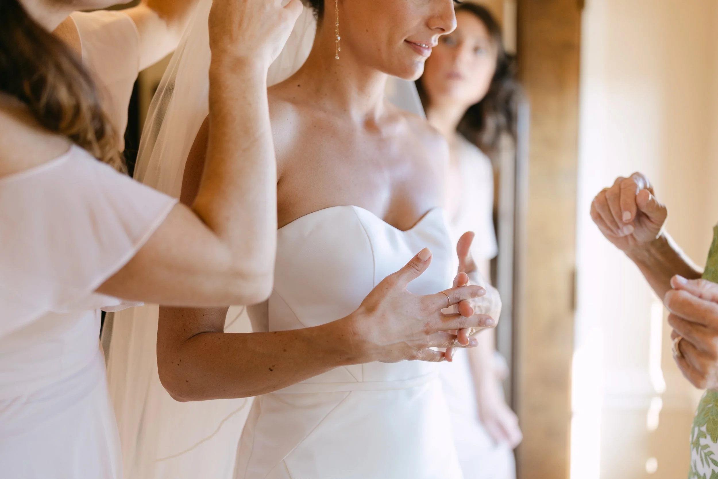 A bride in a white strapless wedding dress with her hands clasped, standing beside bridesmaids during her wedding ceremony, with a person officiating.