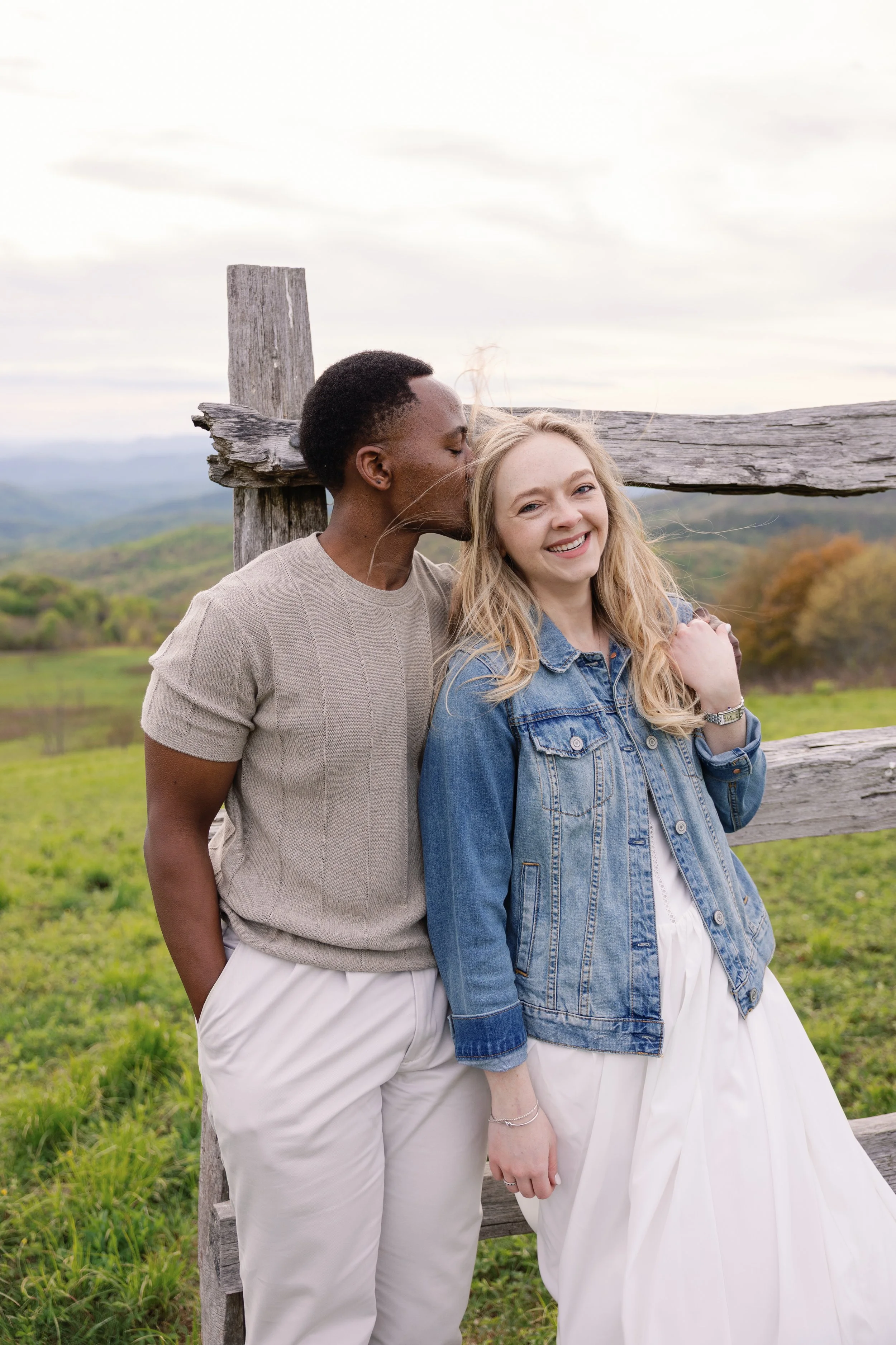 A couple standing outdoors next to a wooden fence, with one kissing the other on the cheek and the woman smiling.