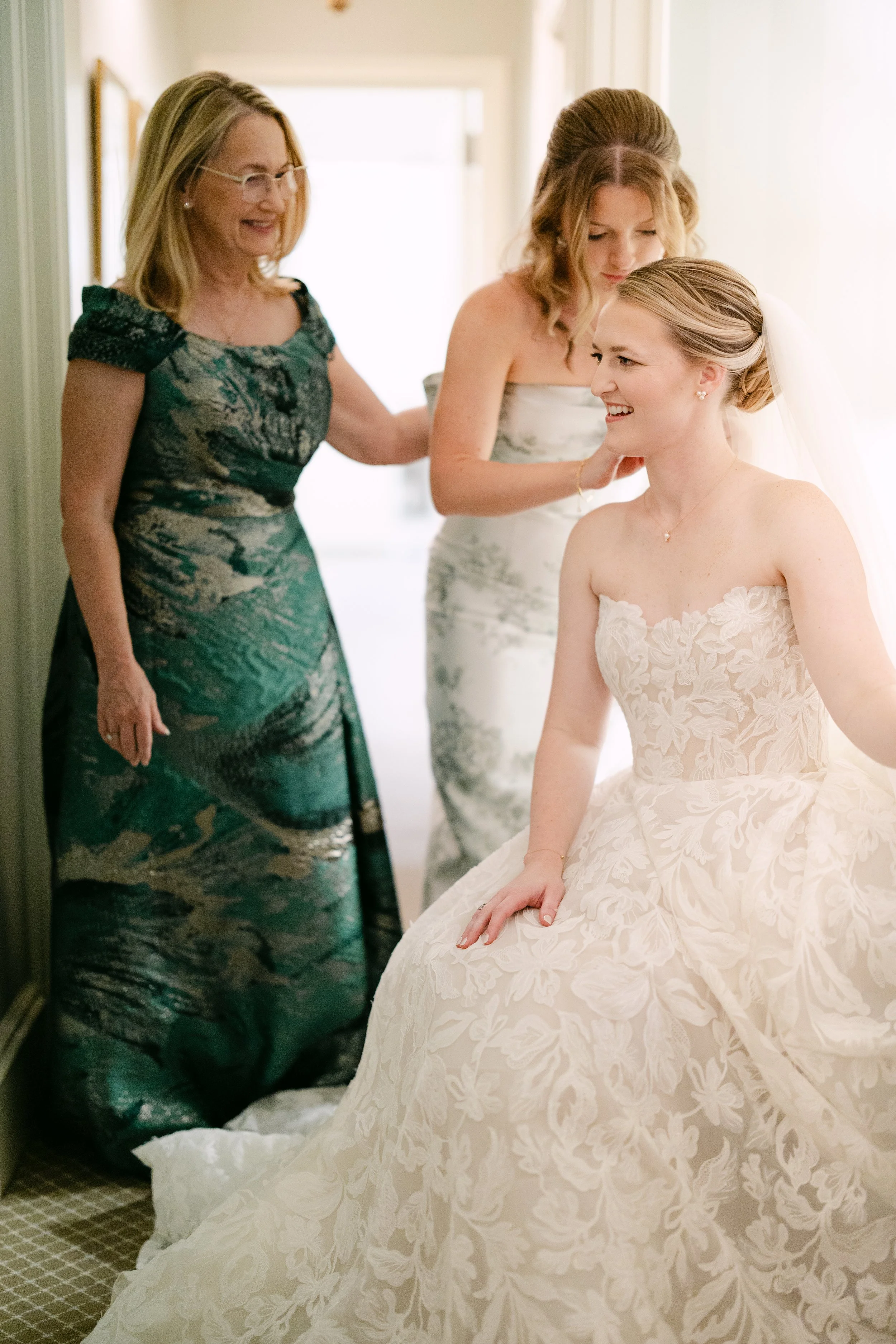 A bride sitting on a chair in a white wedding gown, smiling, with two women, likely her mother and maid of honor, standing behind her and adjusting her hair, in a bright room.