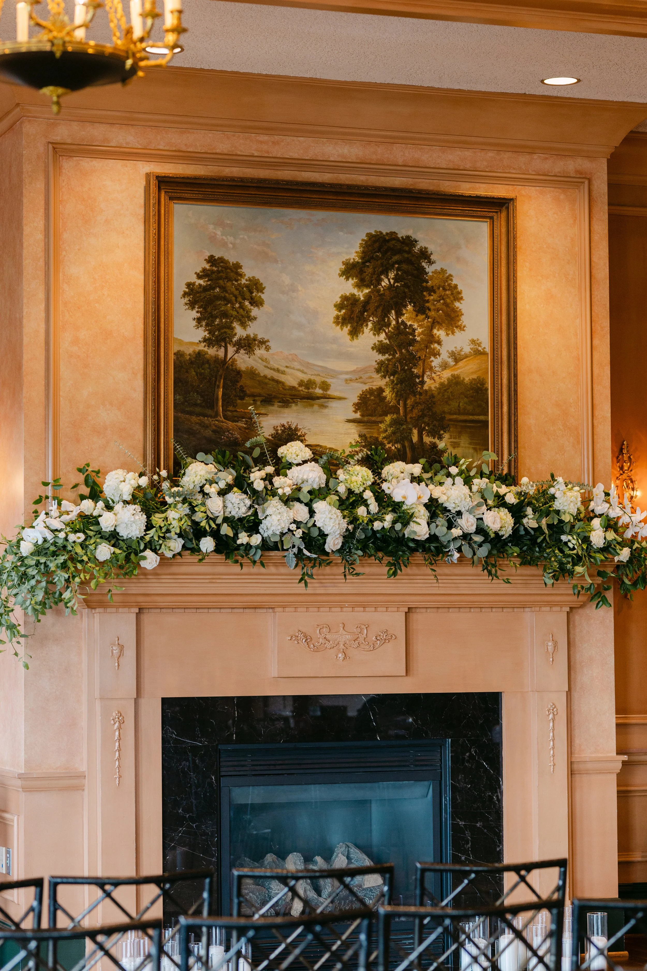 A fireplace decorated with a lush arrangement of white flowers and greenery, with a landscape painting of trees and a river hanging above it.