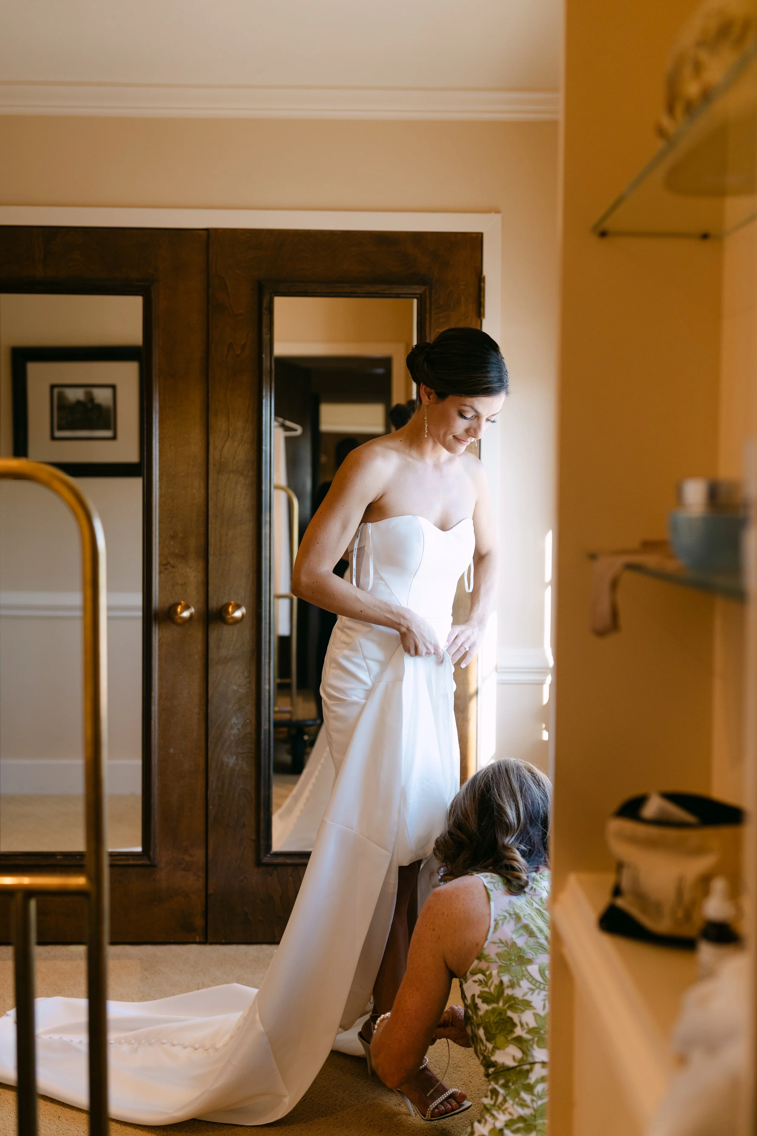A woman in a wedding dress getting ready with help from another woman in a patterned dress, inside a room with warm lighting and wooden furniture.