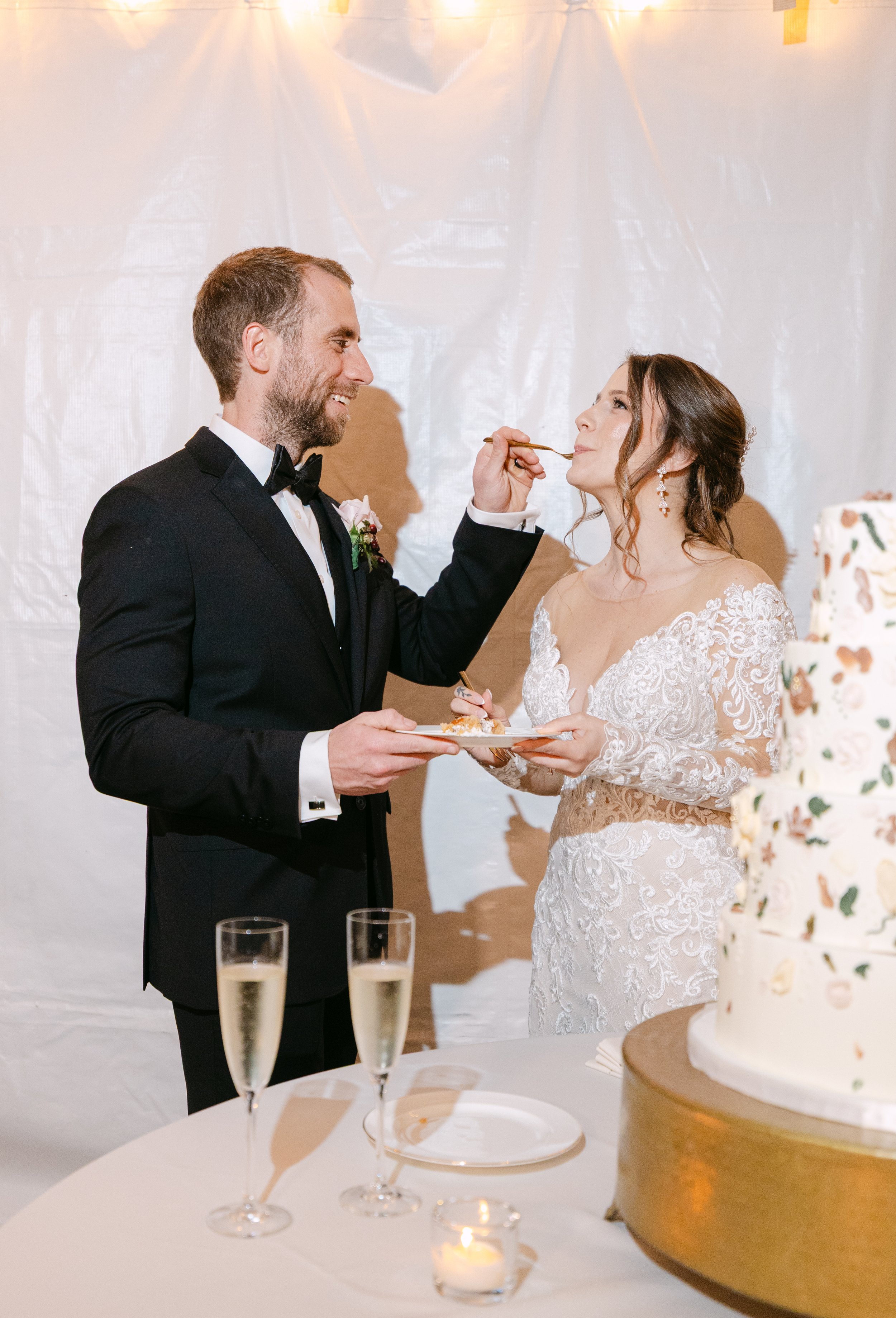 A newlywed couple at their wedding reception sharing a moment as the groom feeds cake to the bride. The bride is wearing a white lace wedding dress and the groom is in a black tuxedo. There is a wedding cake and two glasses of champagne on the table.