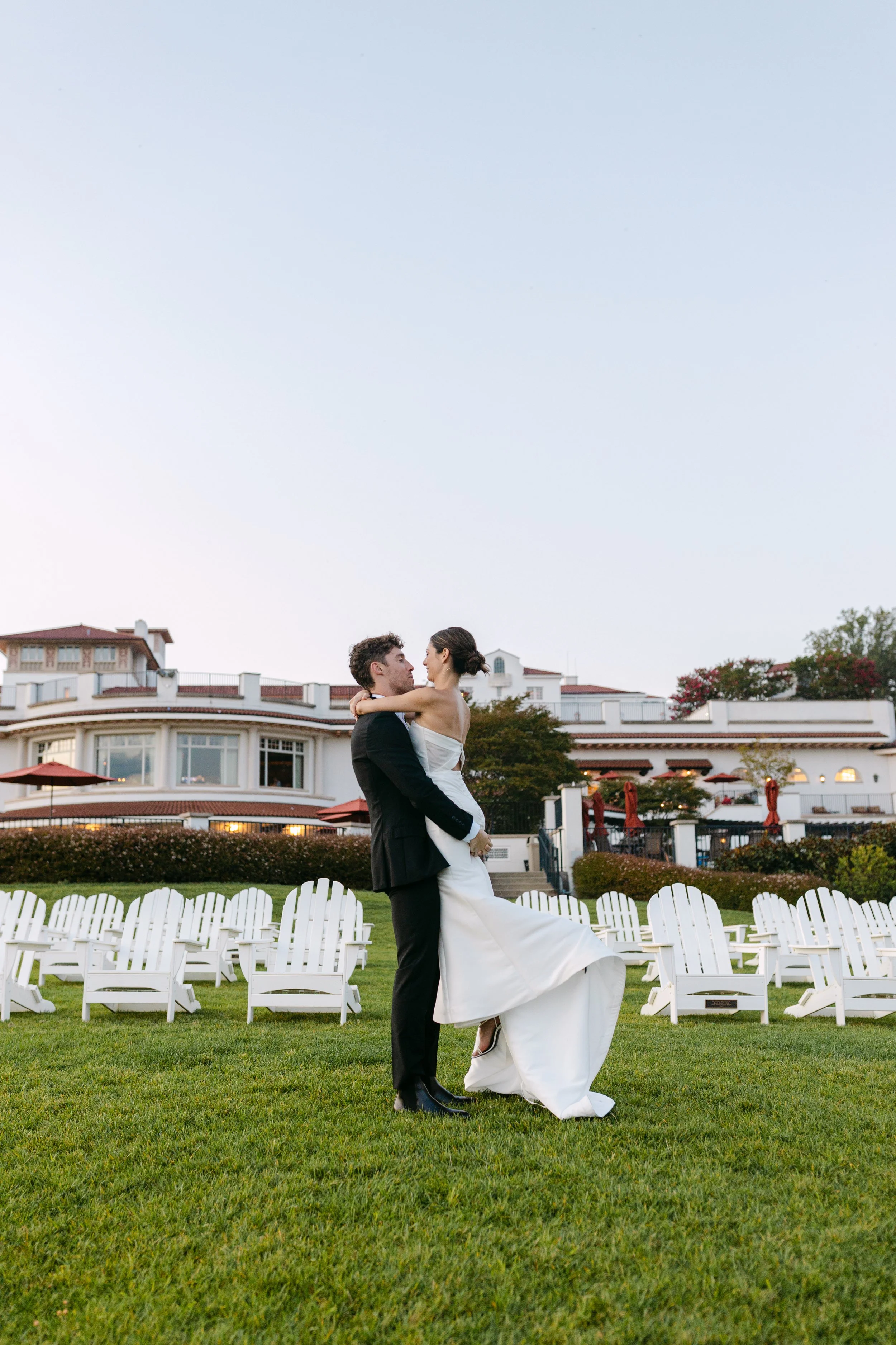 A bride and groom dancing on a lawn with white chairs in the background, during a wedding at a large estate or hotel.