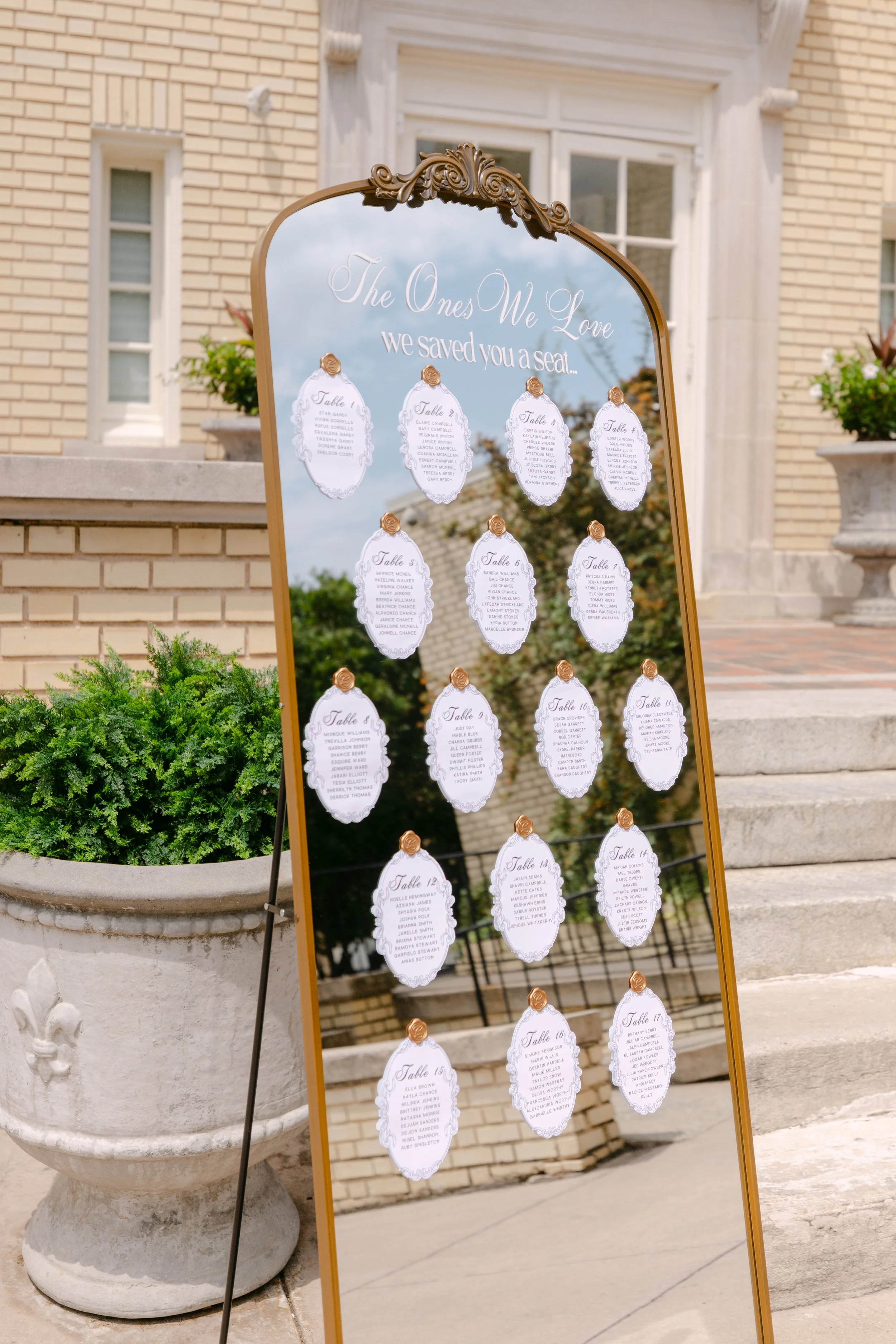 Outdoor wedding seating chart on a standing mirror board displaying table assignments with elegant white and gold design, in front of a brick building and potted plants.