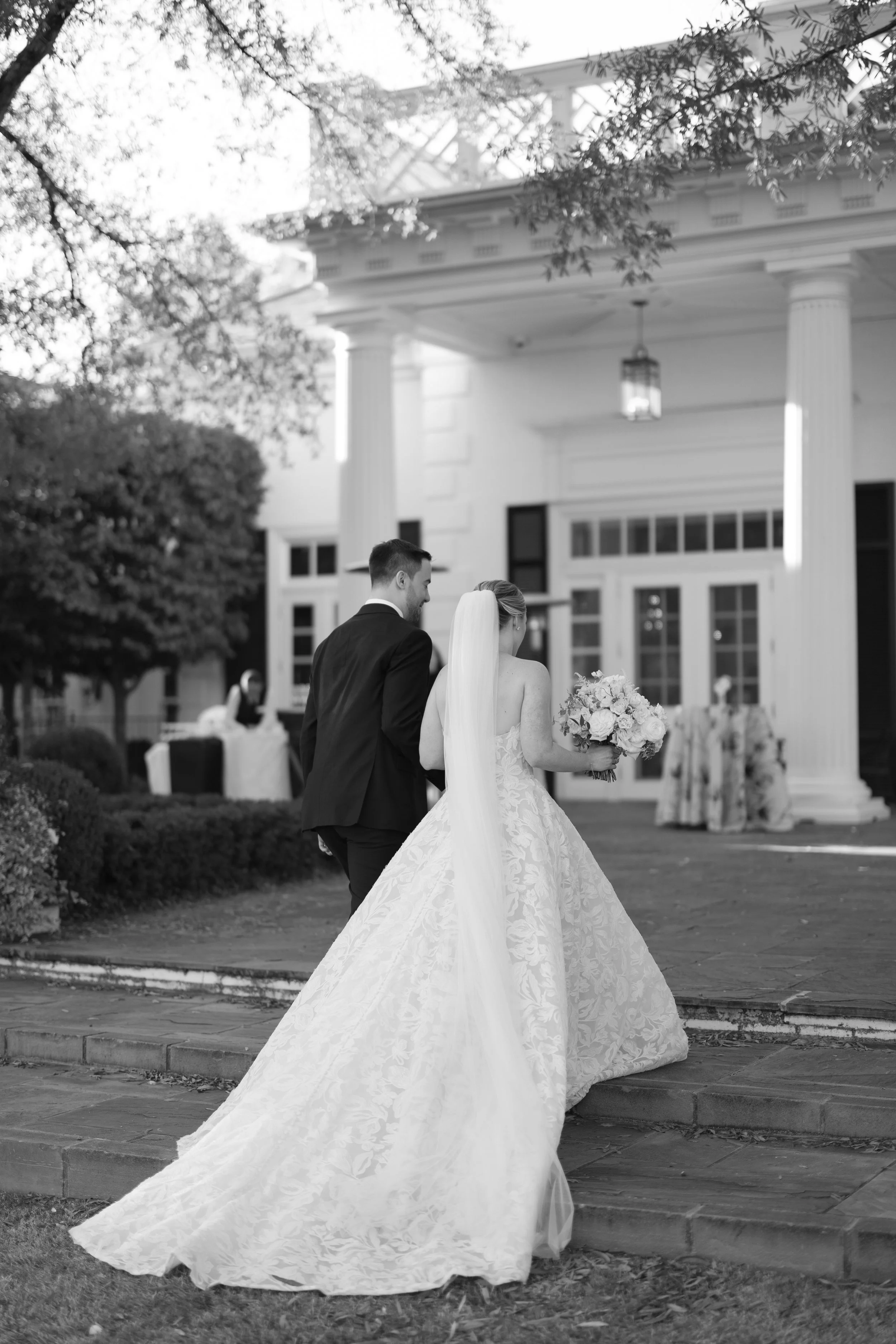 A bride and groom walking up the steps outside a building with columns on a wedding day, with the bride holding a bouquet of flowers.