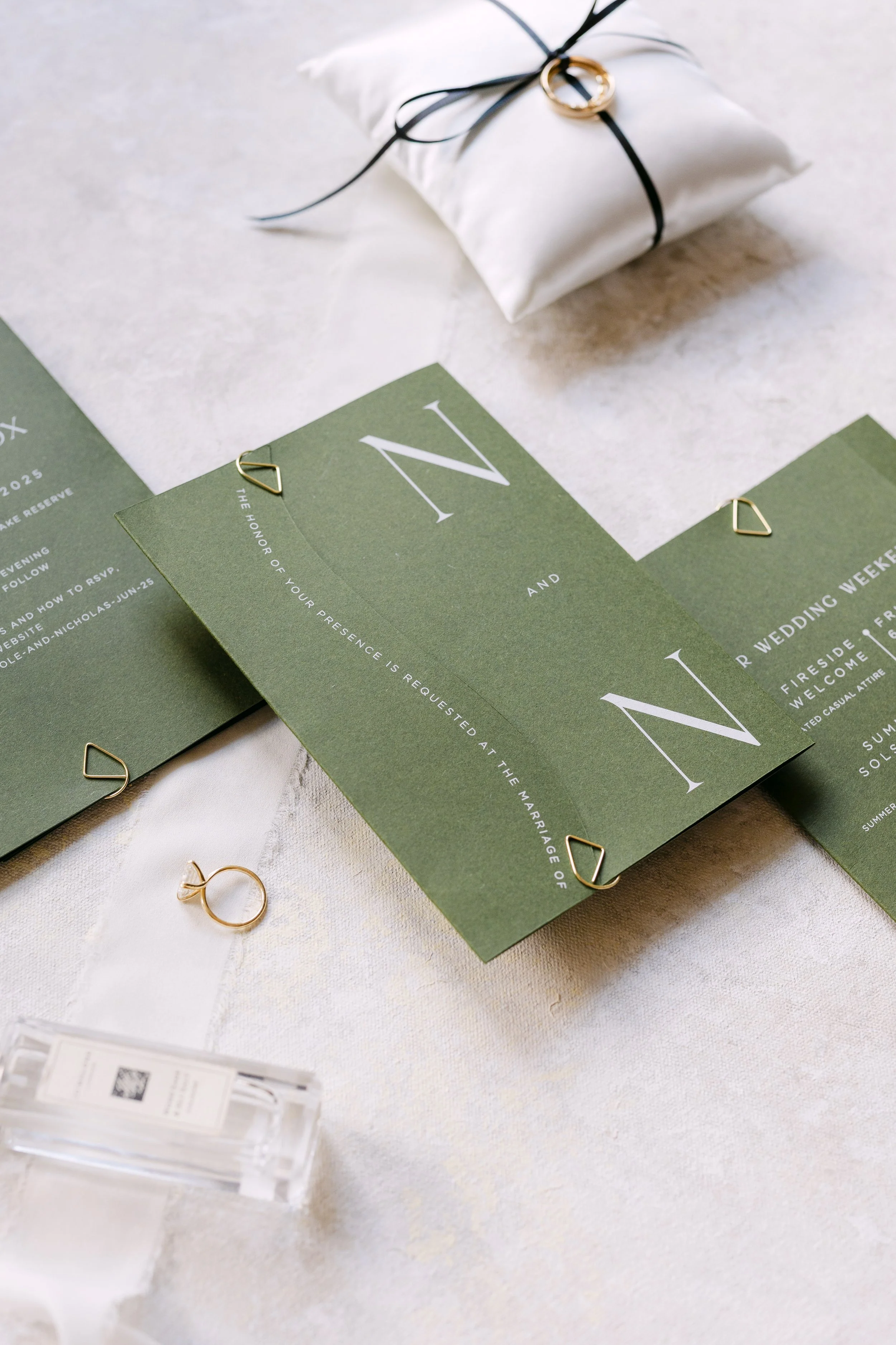 Wedding invitation cards on a white table, some gold rings, a small wrapped gift with black ribbon, and a clear perfume bottle.
