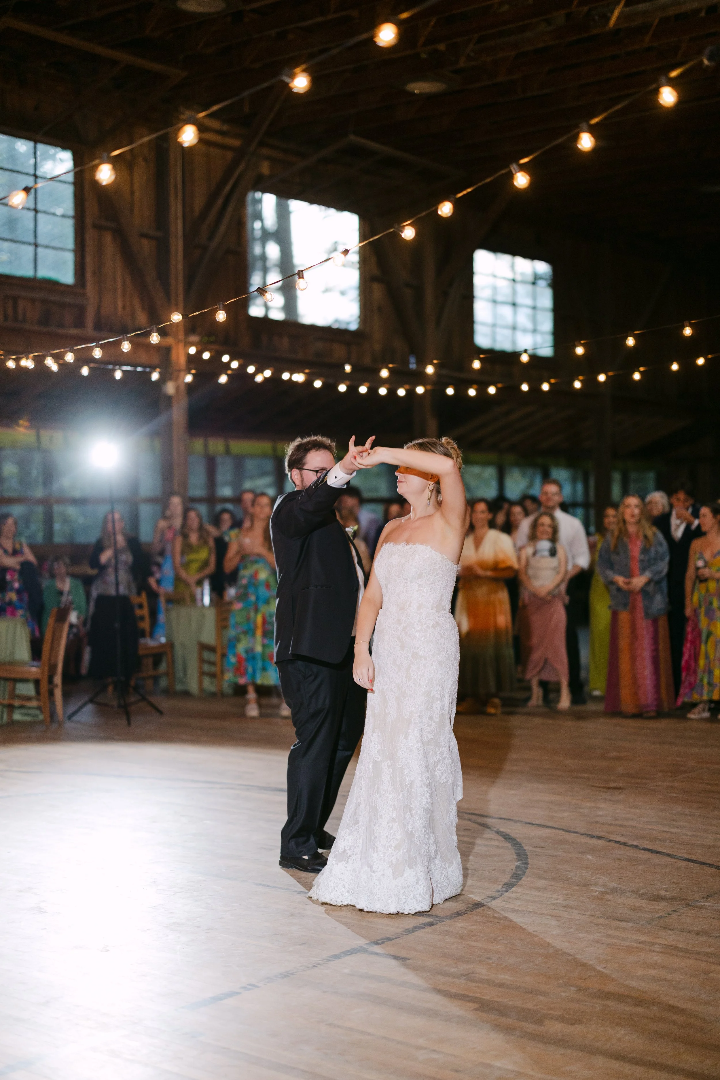 Couple dancing at their wedding reception inside a rustic barn decorated with string lights, with guests watching in the background.