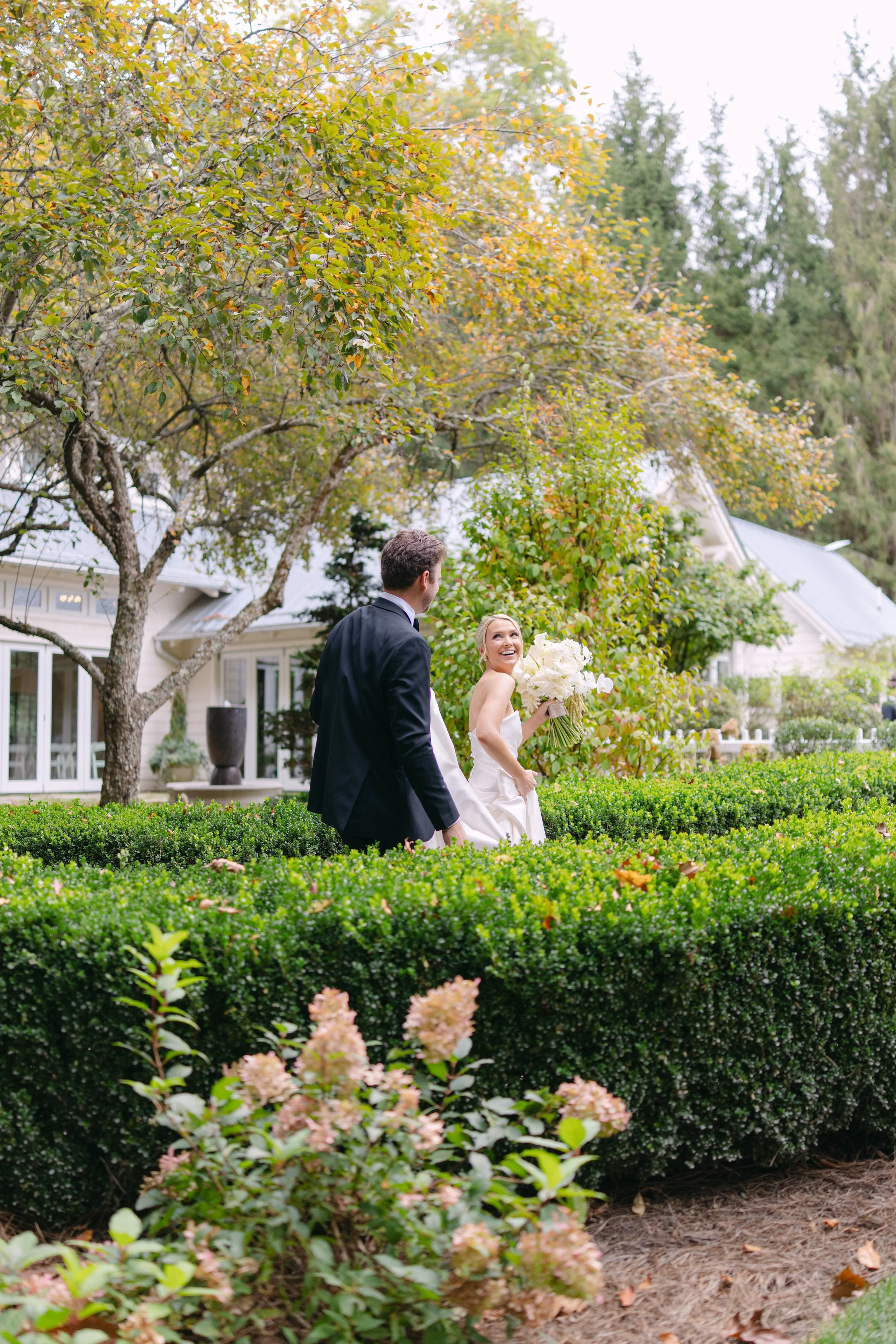Bride holding a white bouquet walks with a groom through a lush garden outside a wedding venue.