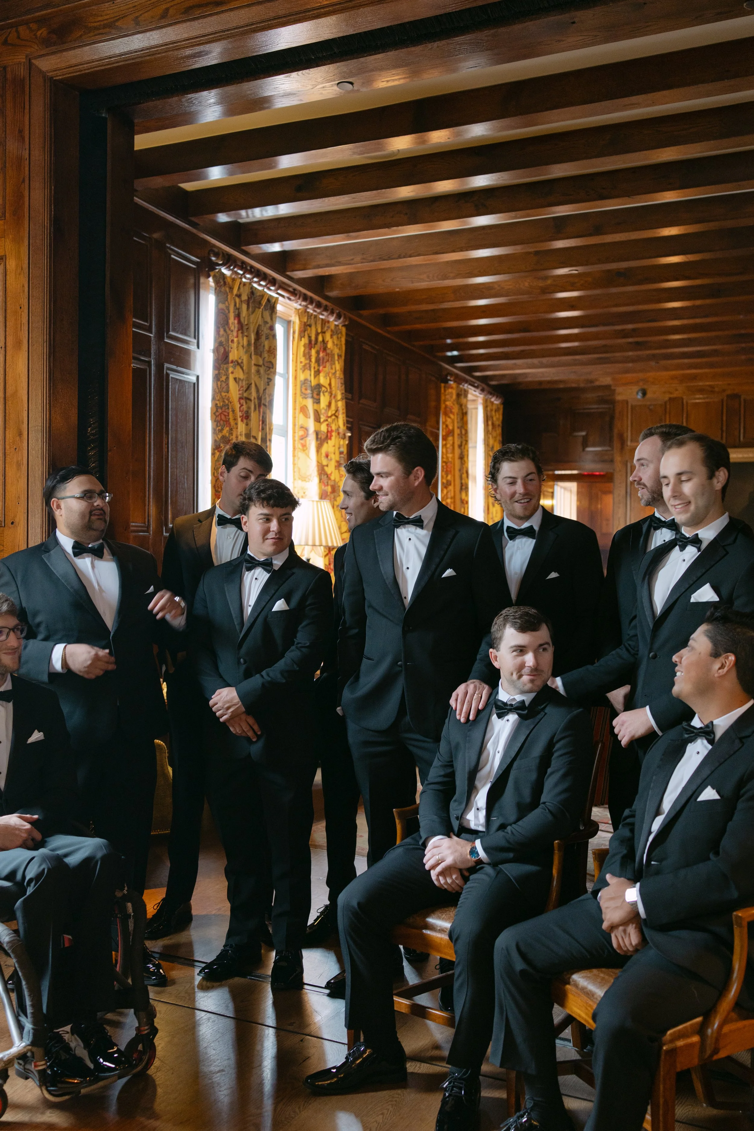 Group of men in black tuxedos and bow ties gathered inside a wood-paneled room.