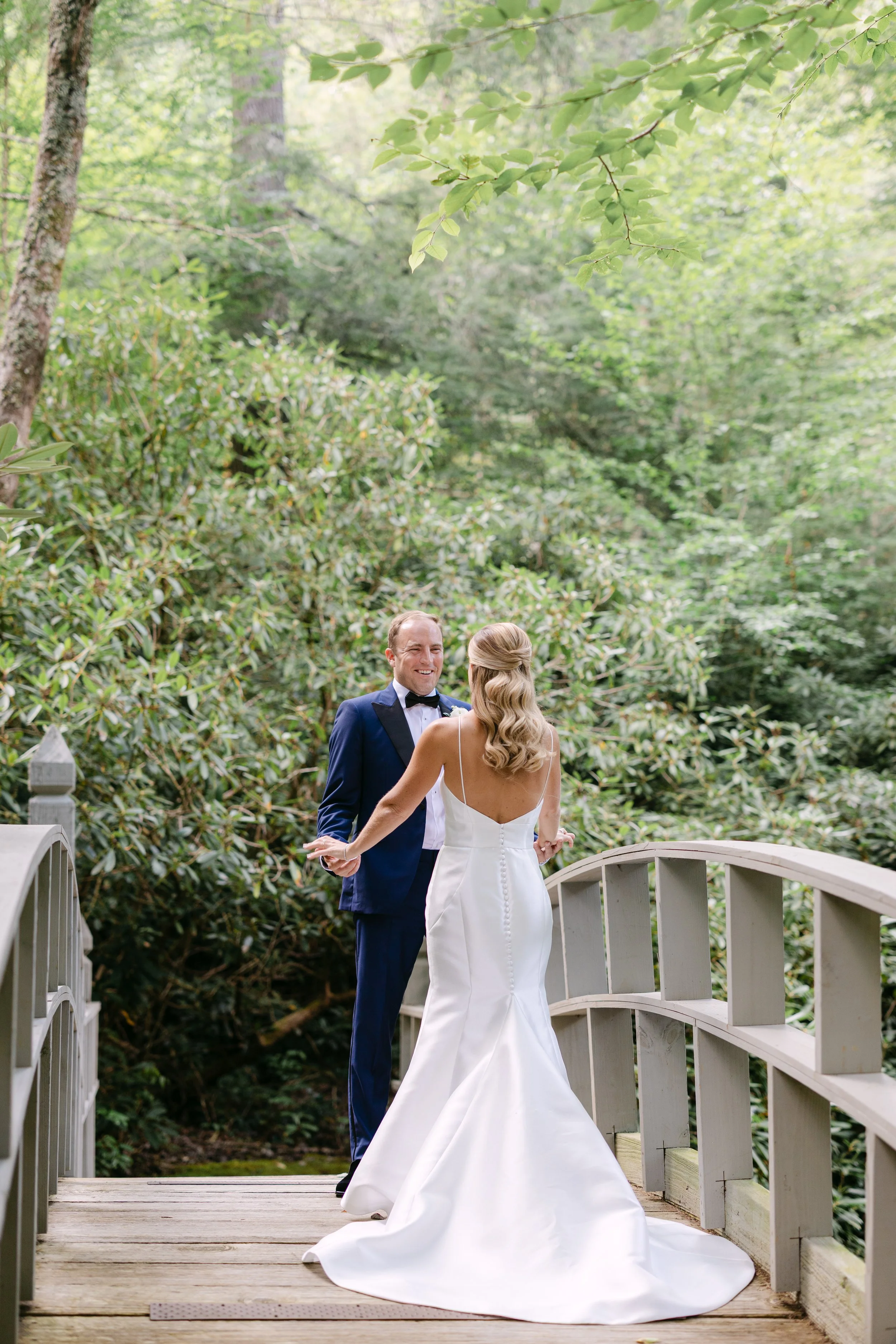 A bride and groom sharing a moment on a wooden bridge surrounded by lush green trees and foliage.