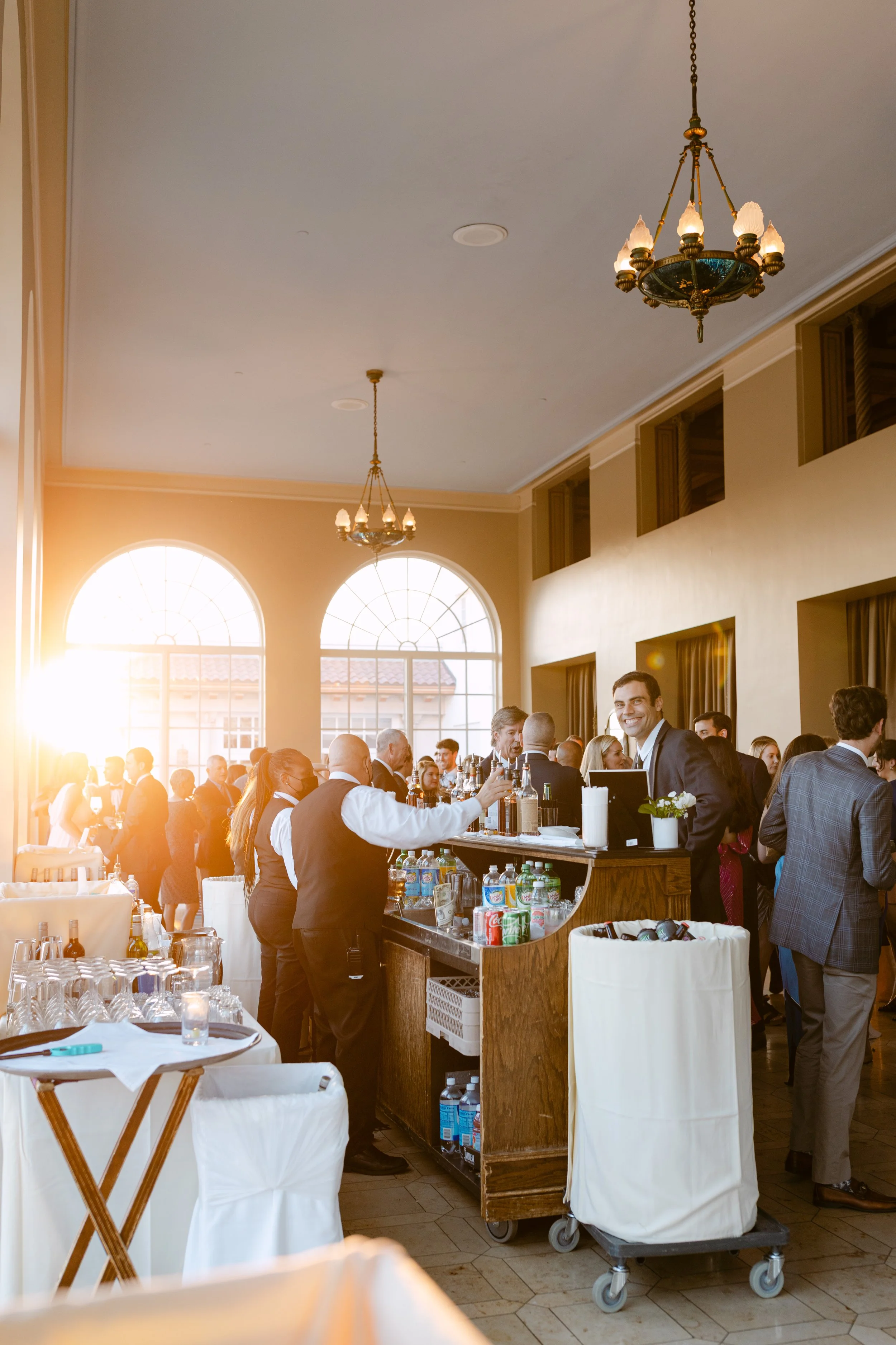 A lively indoor party or wedding reception with people socializing, a bartender serving drinks, and sunlight streaming through large arched windows.