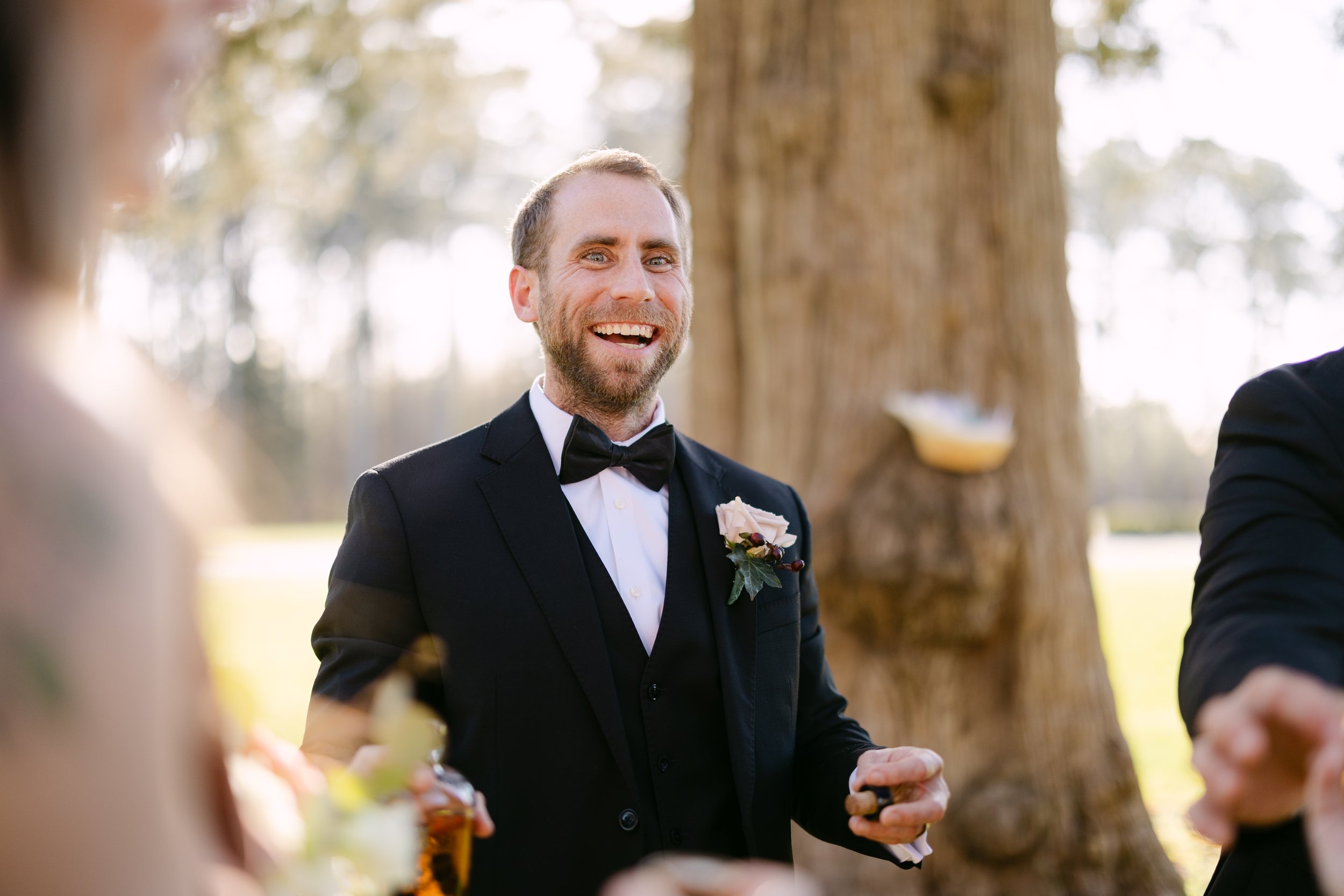 A smiling man in a black tuxedo with a bow tie and a flower boutonniere is at an outdoor event, standing near a large tree, with other people partially visible around him.