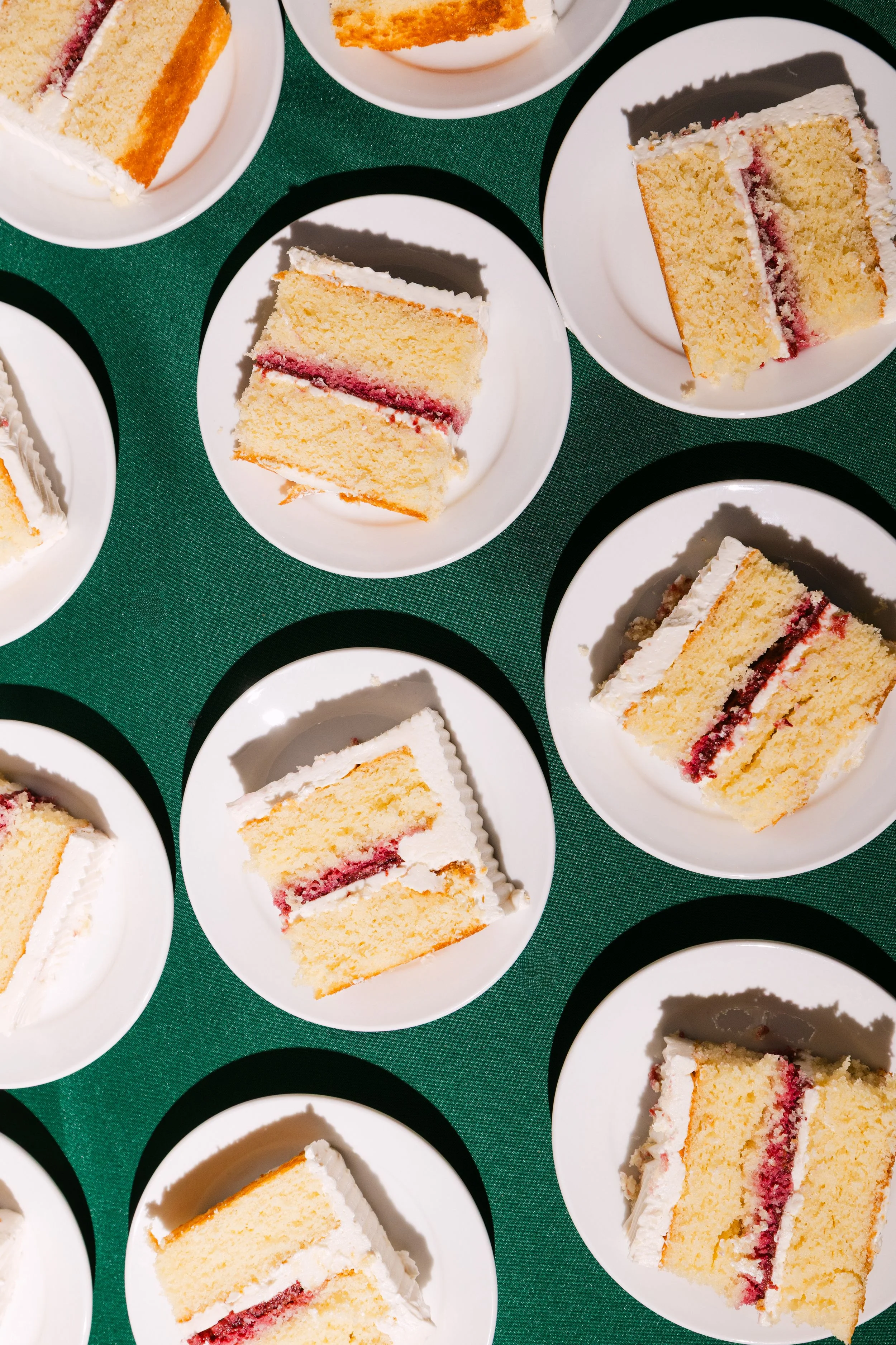 Multiple slices of birthday cake with white frosting and red jam filling served on white plates on a green table.